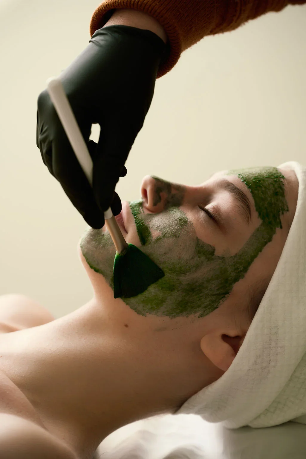 A person with a towel wrapped around their head receives a facial treatment with green mask applying on their face while lying down, and a professional wearing gloves uses a brush to apply the mask.