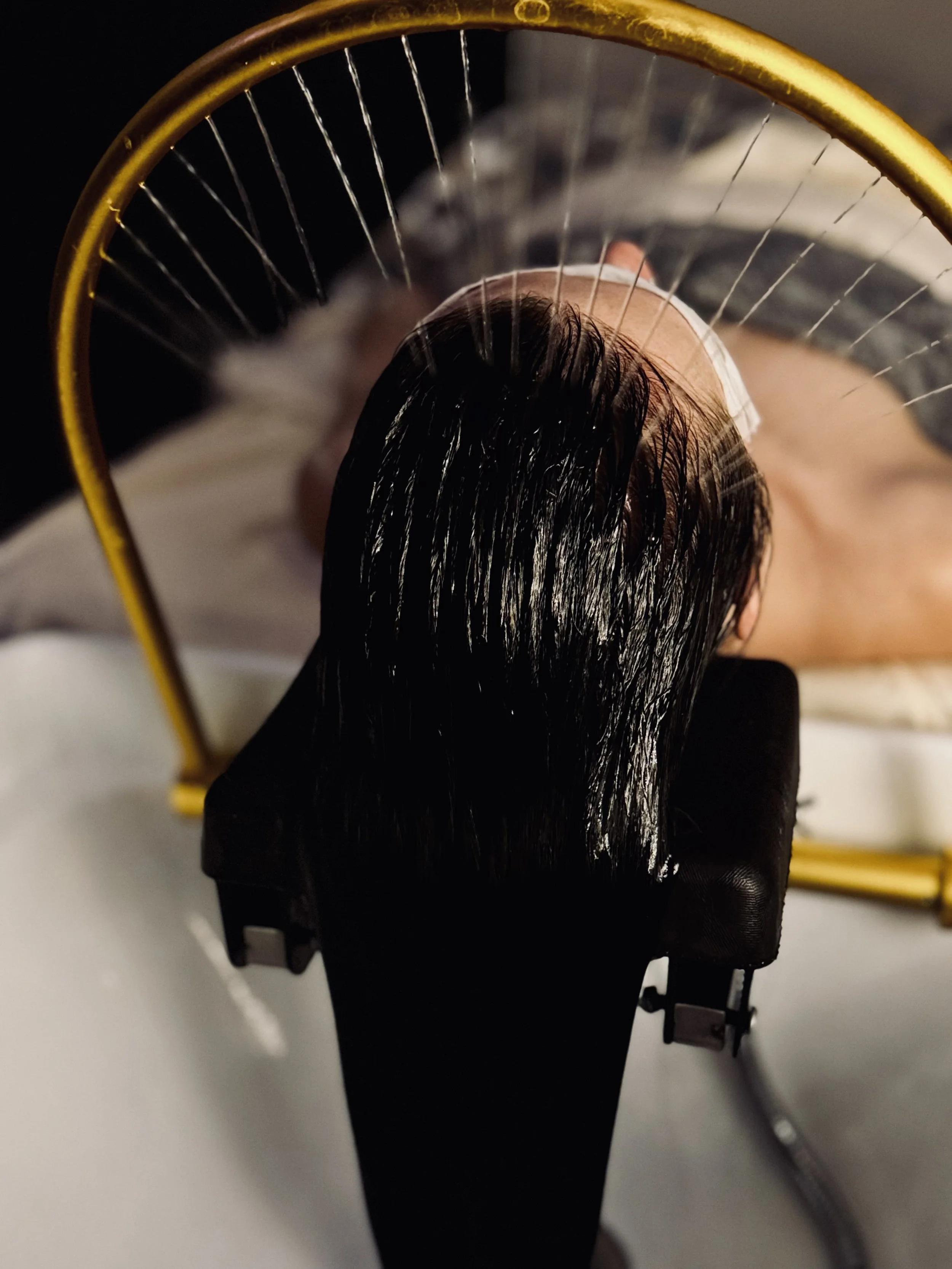 Top view of a person with wet, dark hair lying in a reclined medical or spa chair with a protective eye mask on, and a yellow circular frame in the background.