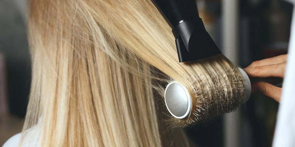 A person with blonde hair uses a round curling iron or hot roller to style their hair.