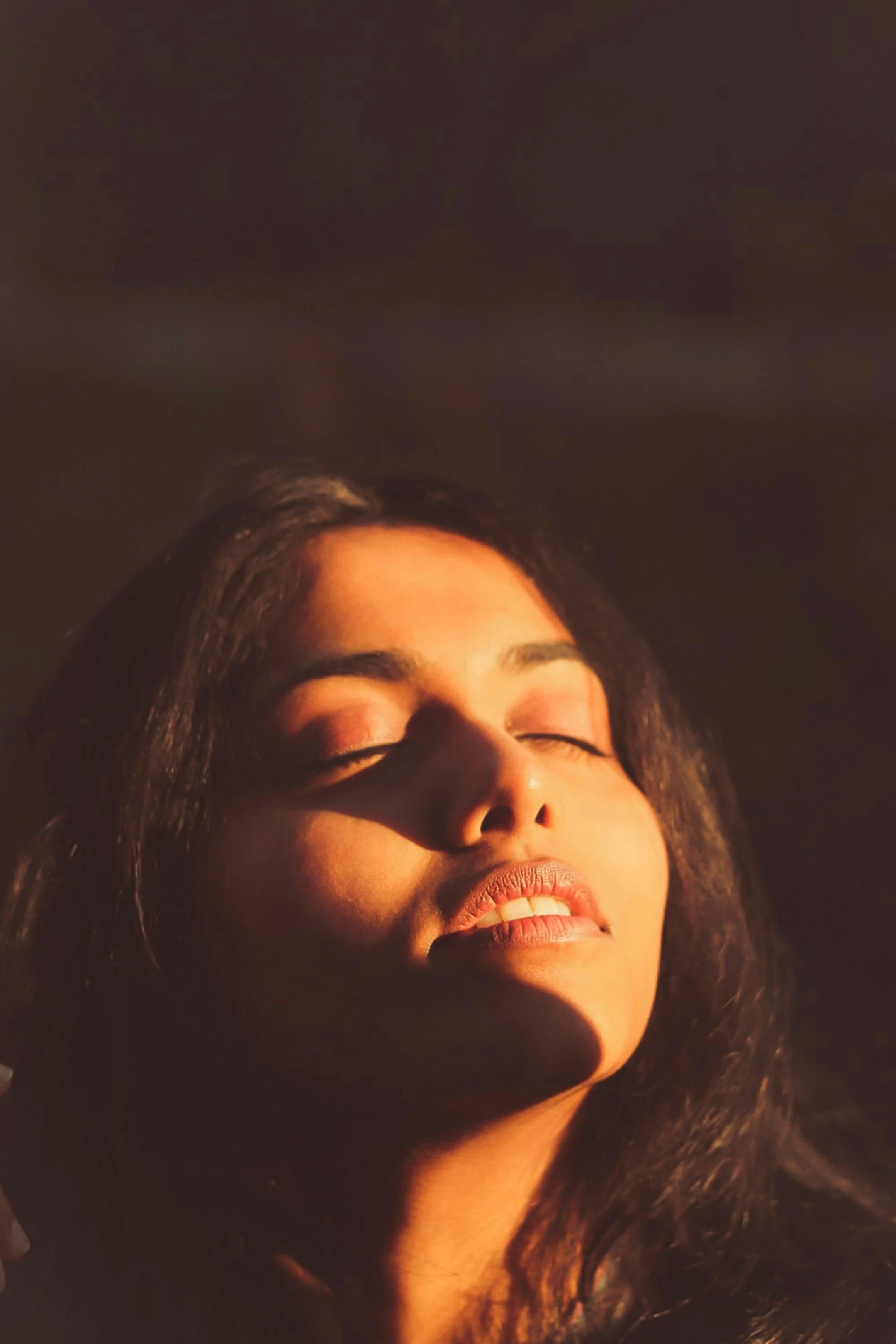 Close-up of a woman's face with closed eyes and a smile, bathed in warm light, dark background.