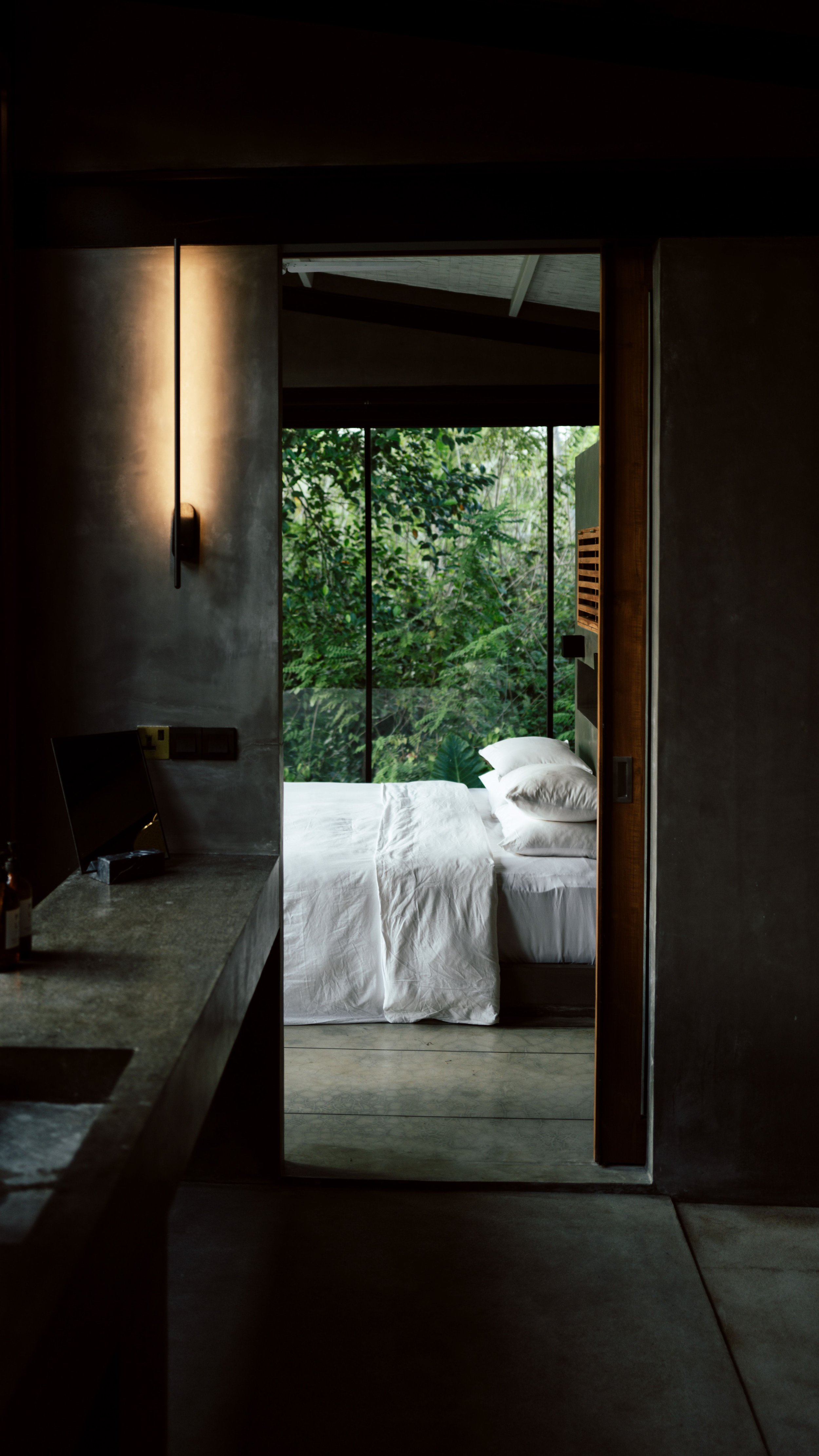 A bedroom with a bed covered in white bedding and pillows, positioned by large windows looking out into lush green foliage outside. The room has minimalistic decor with a concrete wall and a wall-mounted lamp.