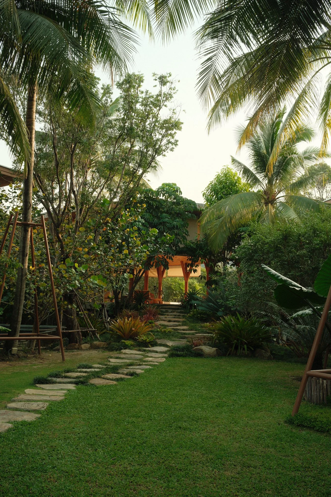 A lush tropical garden with a stone pathway leading to a wooden gazebo, surrounded by various green plants, trees, and palm trees under a bright sky.