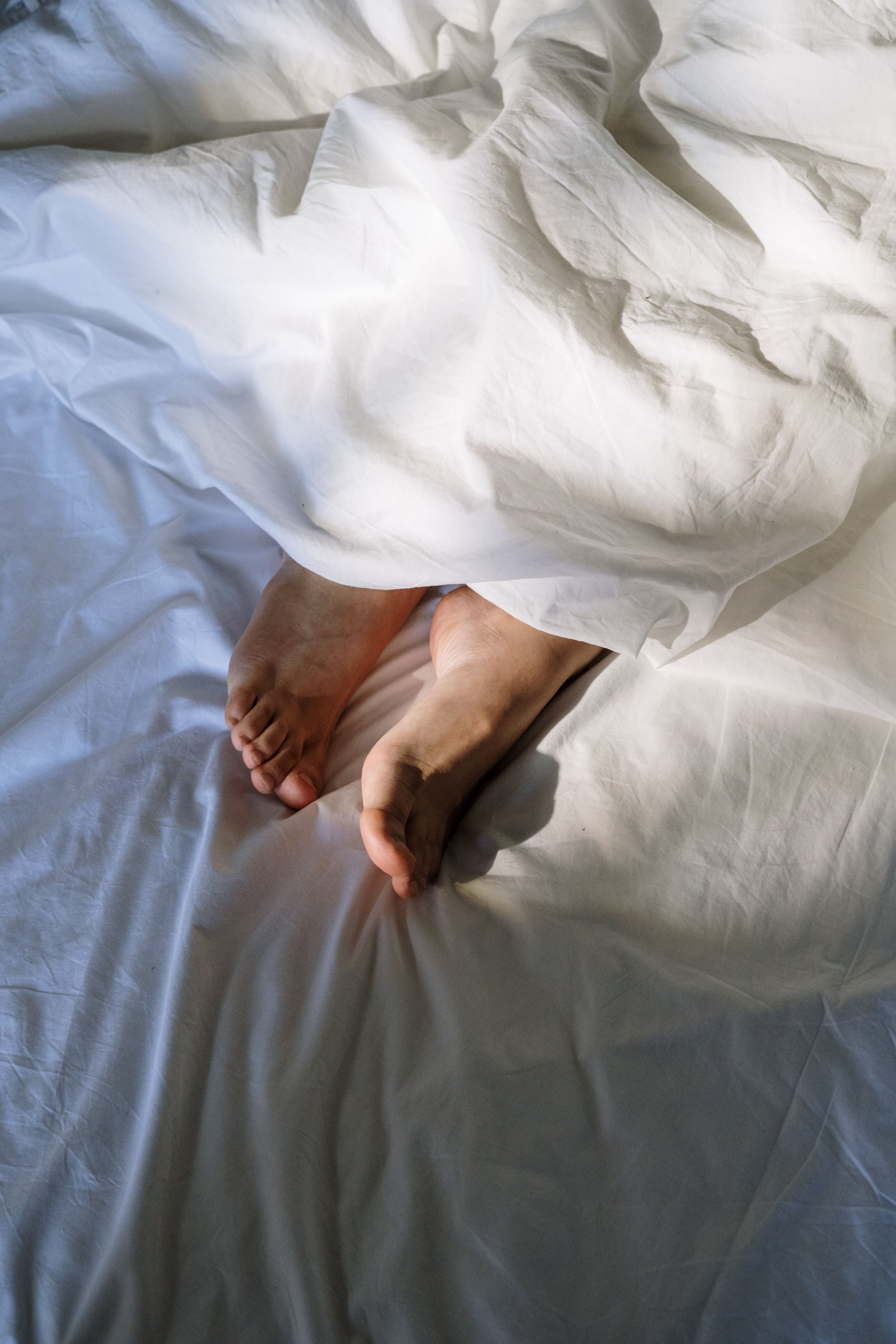 A person lying in bed with pale skin and bare feet exposed, covered by a white sheet.