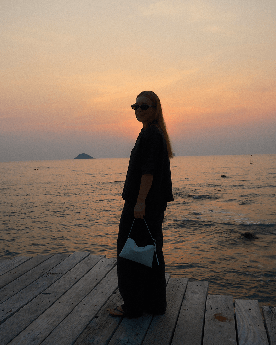 On the picture you see me, a young woman wearing sunglasses. In the back you see the open water and the sun is about to go down.