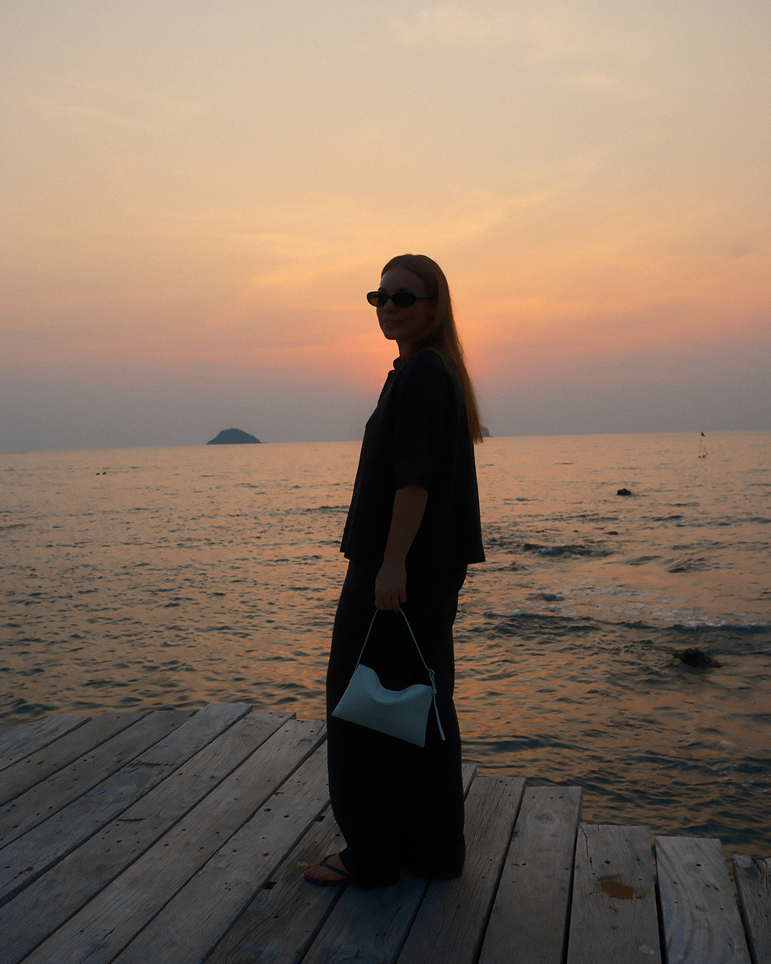 A woman standing on a wooden pier by the ocean at sunset, wearing sunglasses, black outfit, holding a white purse.