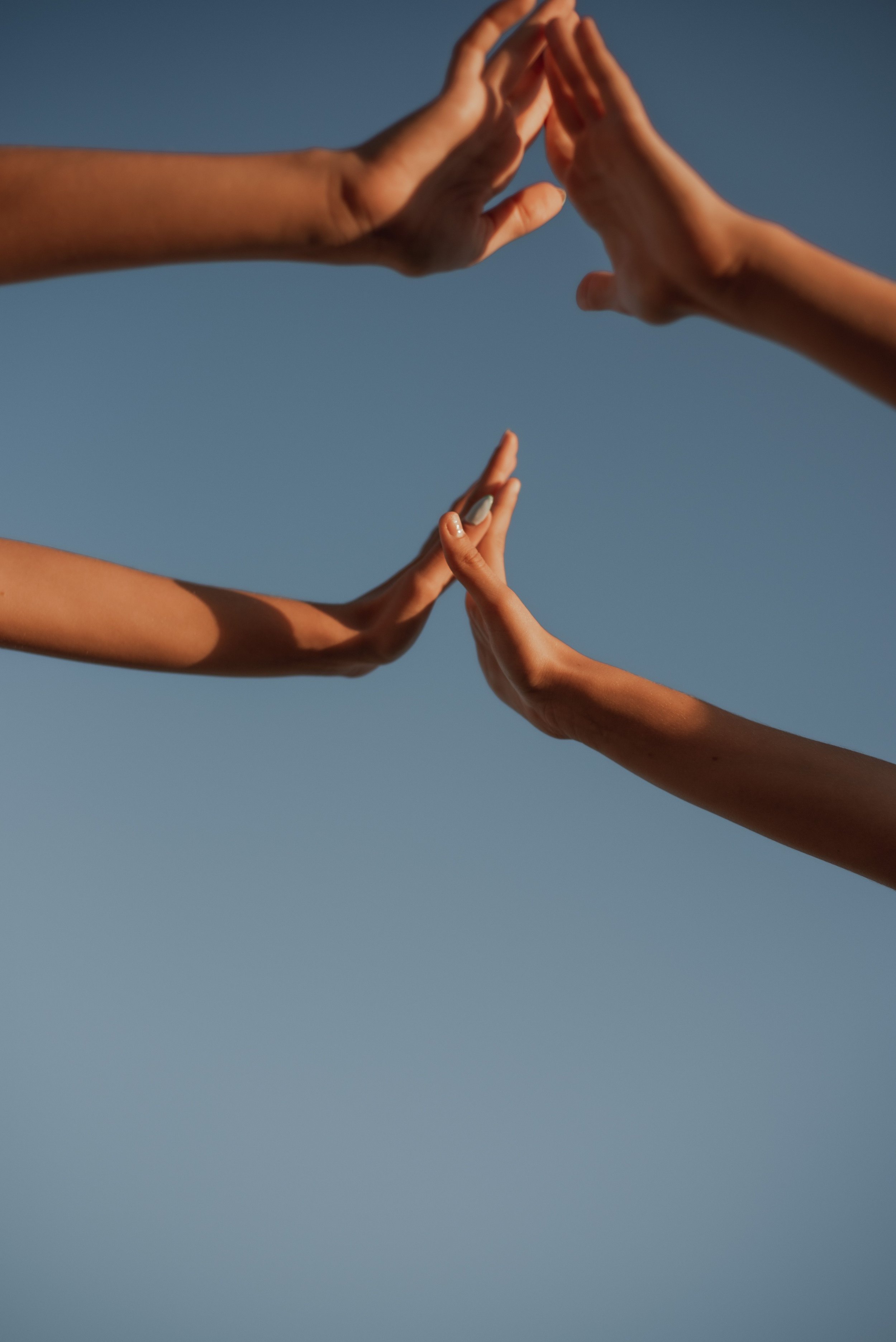 Two pairs of hands reaching towards each other and forming a triangle shape against a clear blue sky.