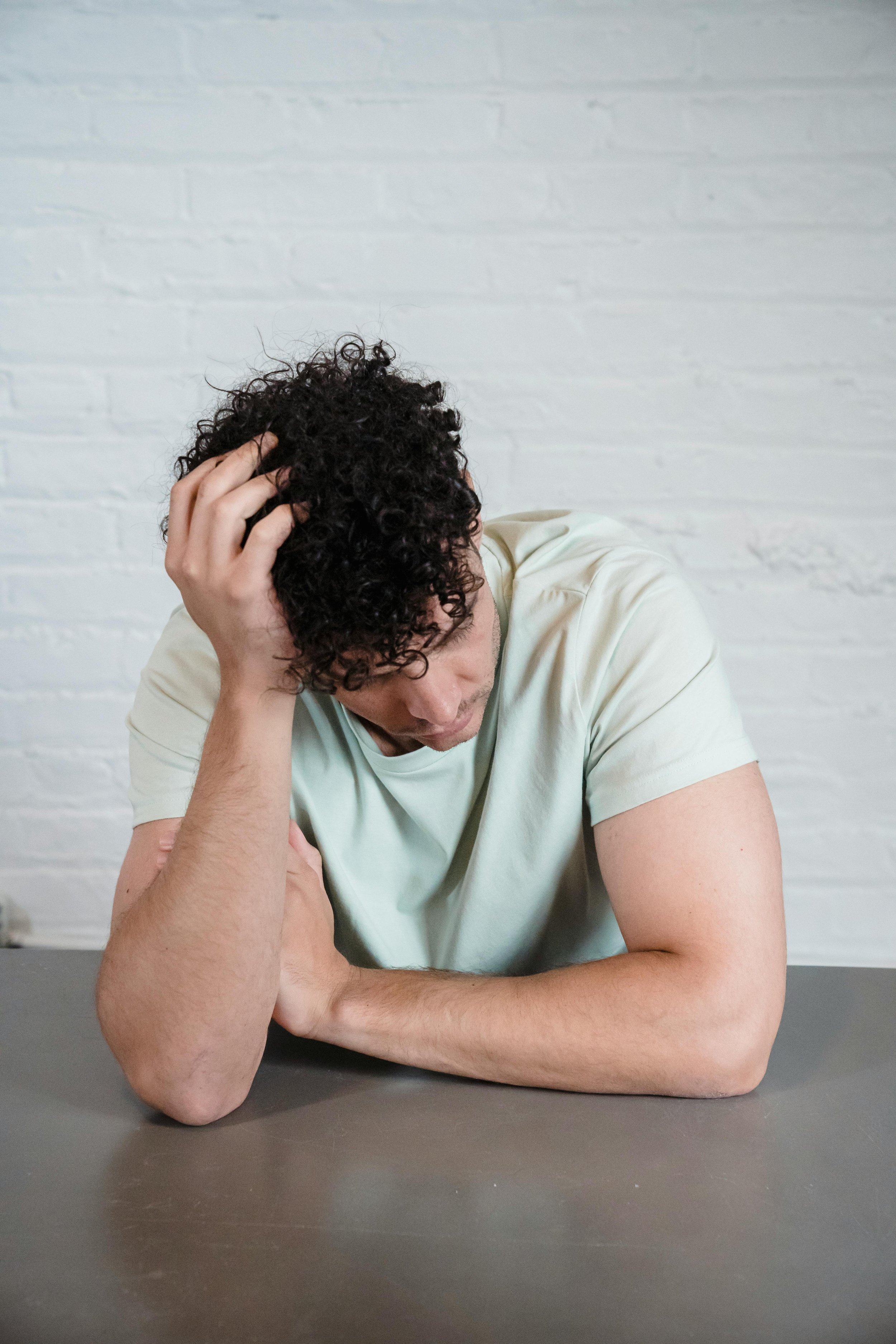 A man with curly dark hair sitting at a table, resting his head on his left hand with his eyes closed, appearing distressed or contemplative.