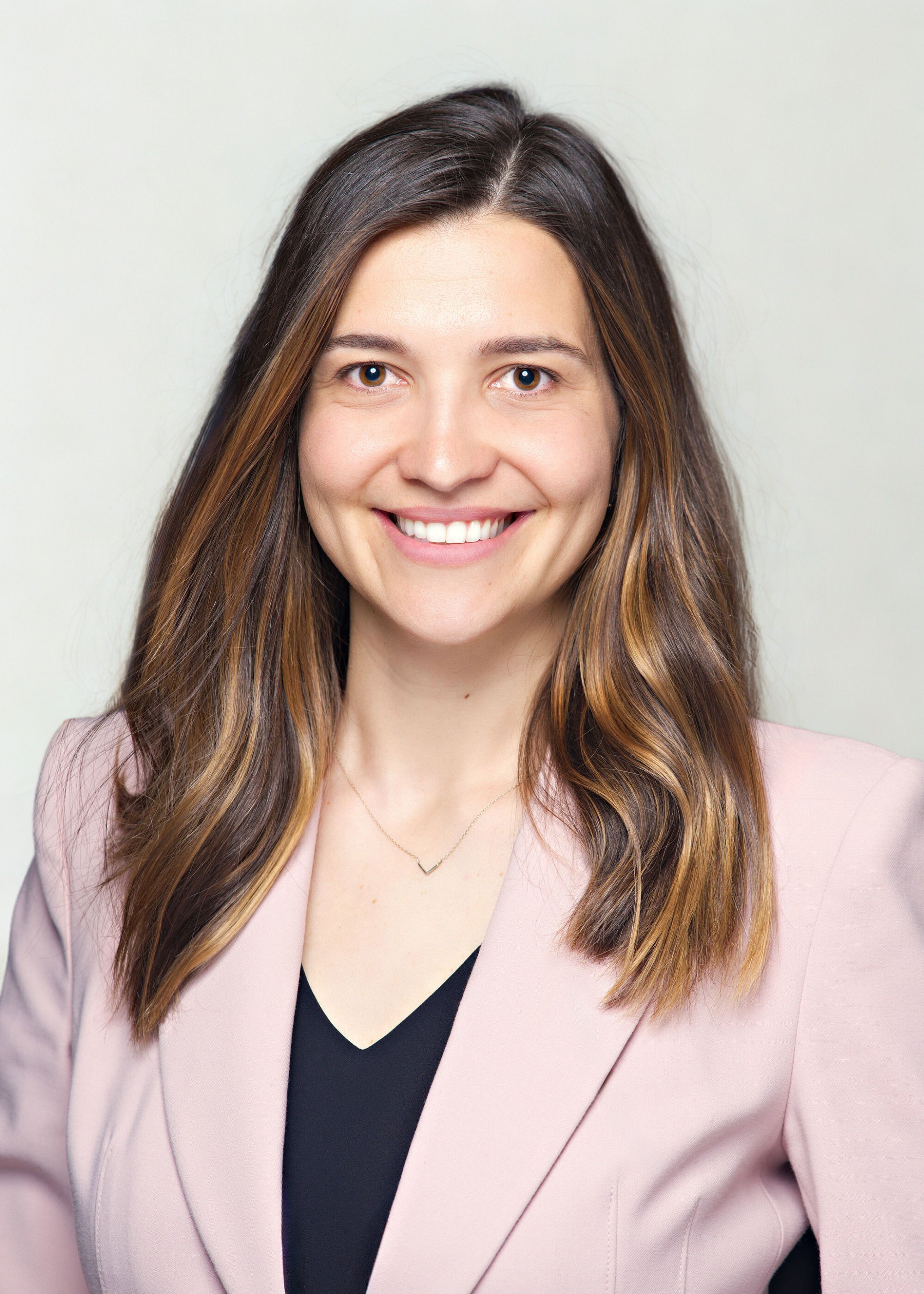 Portrait of a smiling woman with long brown hair, wearing a light pink blazer and a black top, against a plain background.
