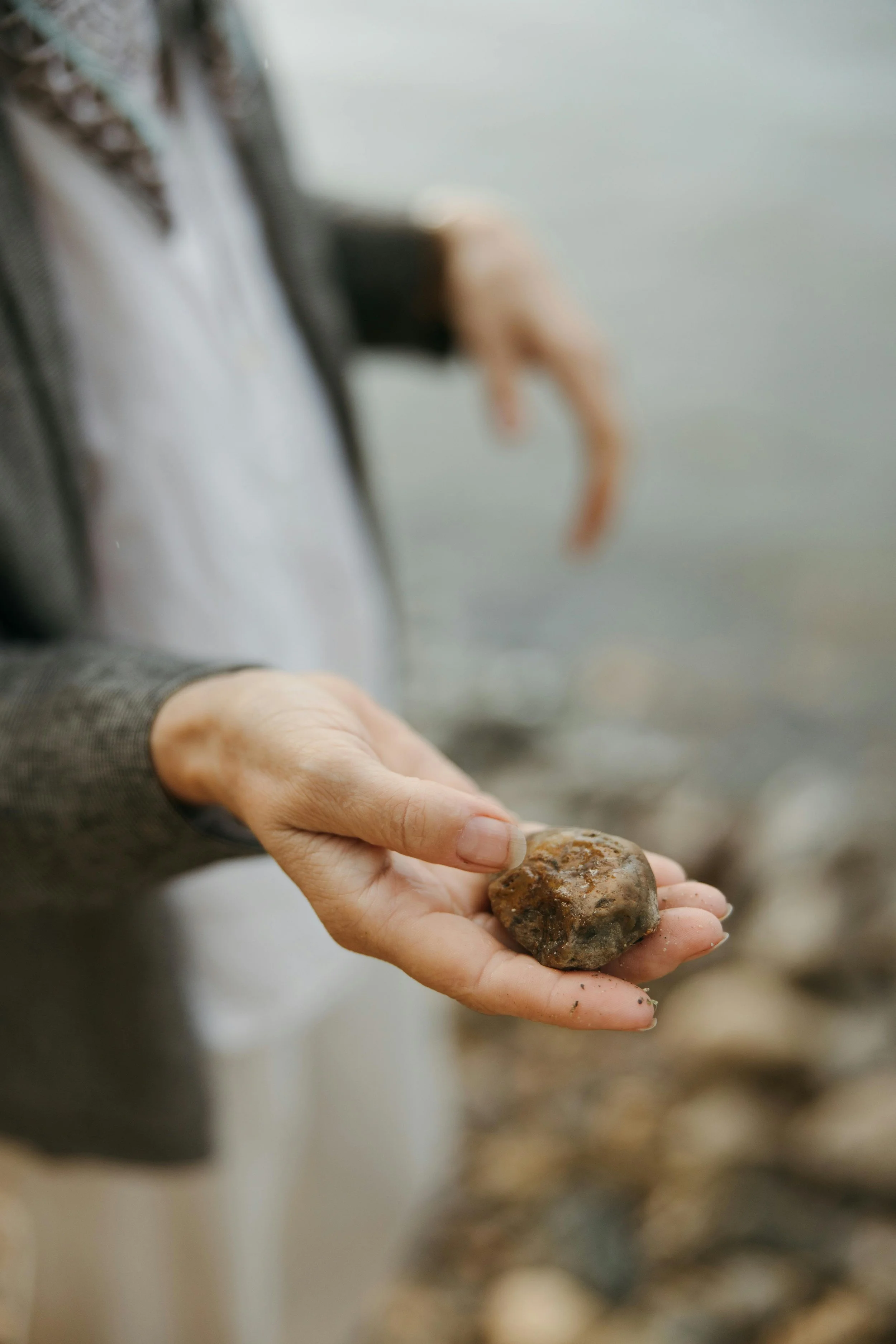 A person holding a small, rounded stone in their hand near a body of water with a blurry background of more rocks.