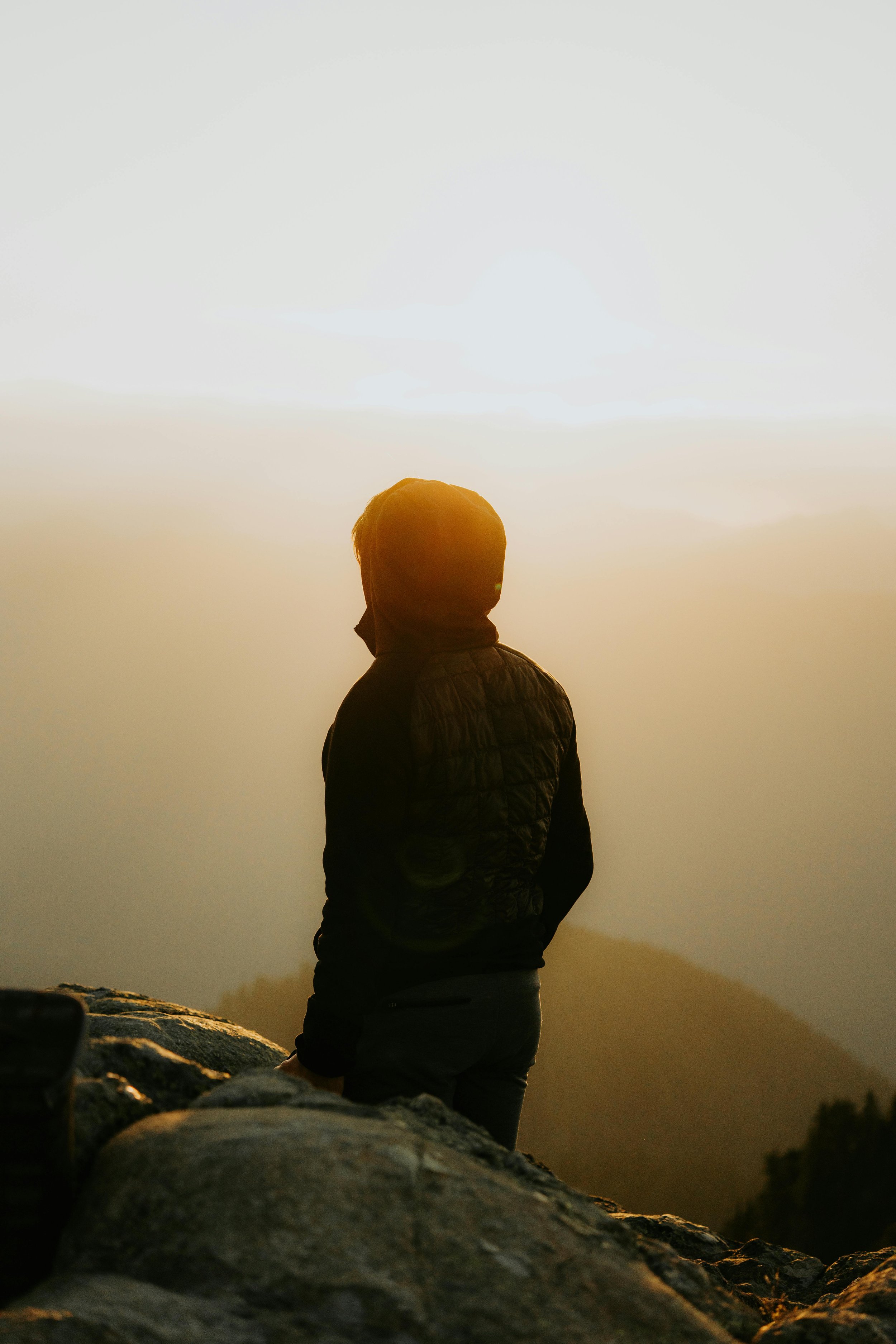 A person standing on rocky terrain, gazing at a sunset or sunrise over a mountainous landscape with a cloudy sky.