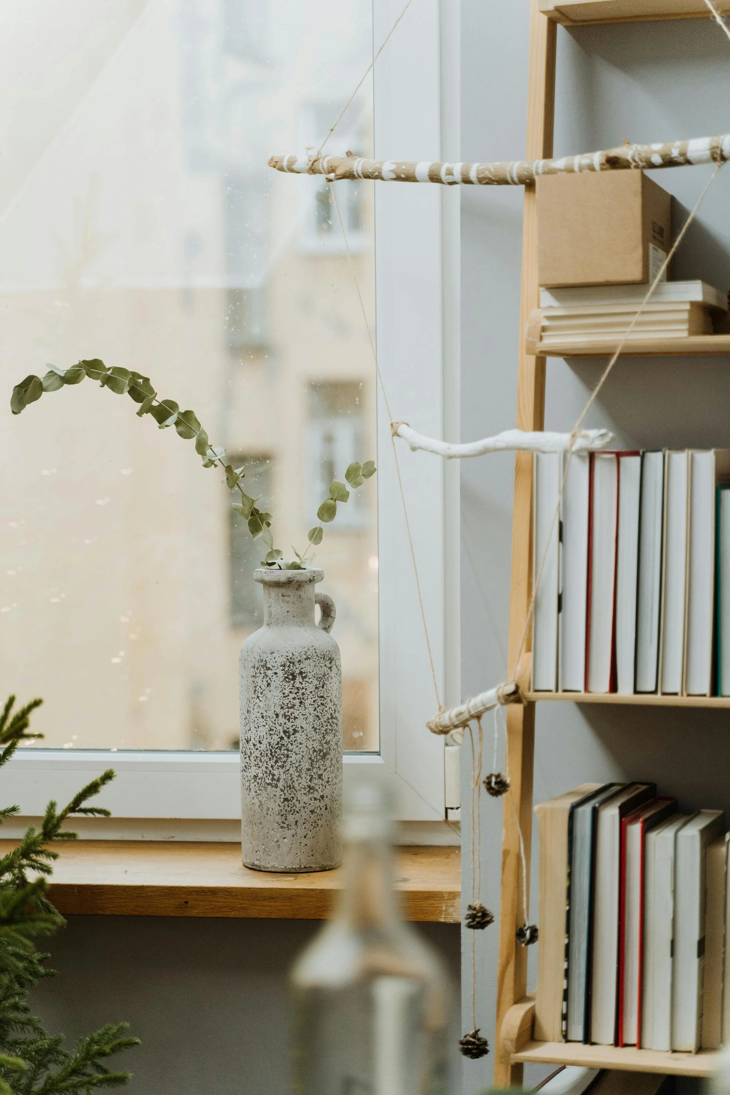 A small, unfinished branch-shaped wooden photo display holder hanging with string on a window, with a weathered white ceramic vase with a small branch with green leaves on a wooden window sill, next to a bookshelf with books.