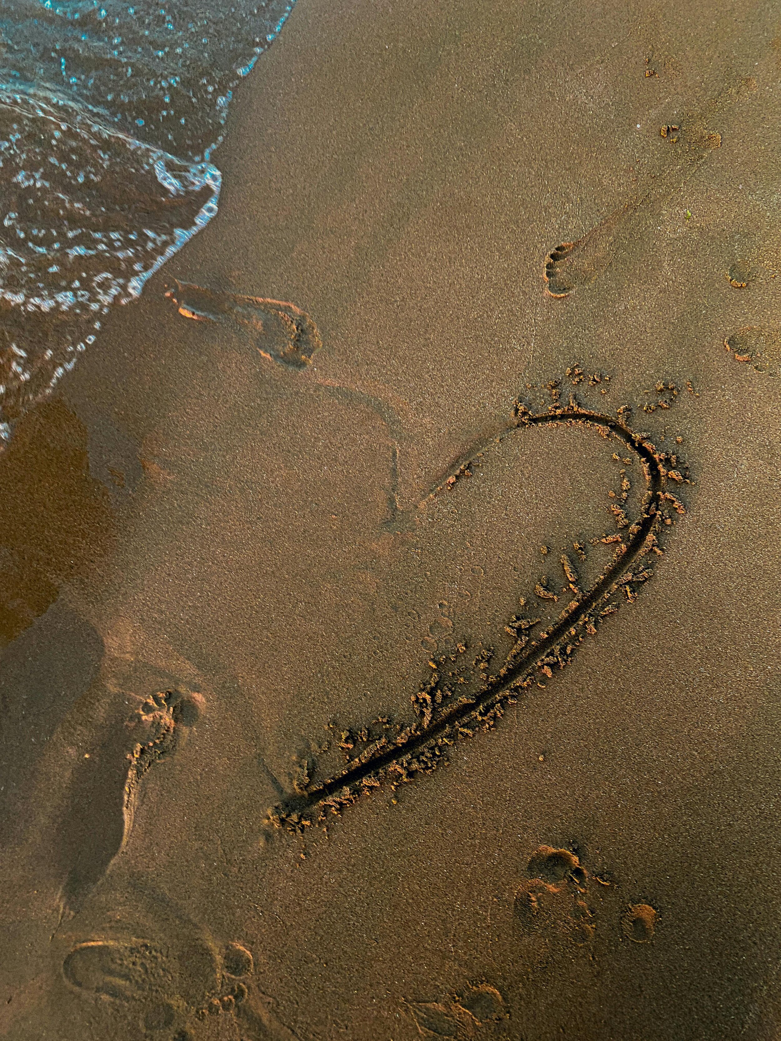 Footprints and a heart drawn with sand on the beach, with a wave approaching.
