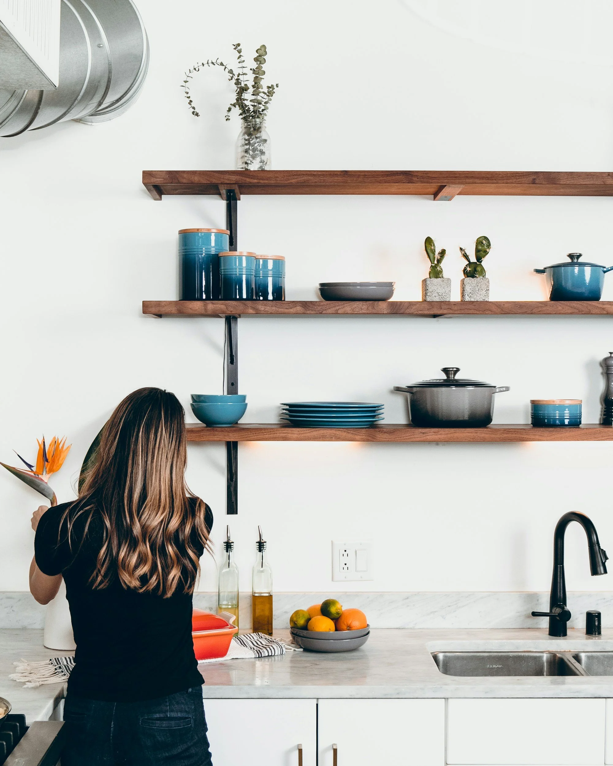 A woman with long wavy hair standing in a modern kitchen, holding a flower in her hand near the counter with a bowl of fruit and two bottles of oil or vinegar. Open wooden shelves with dishes, bowls, plants, and pots are mounted on the white wall above the counter. A black faucet and sink are visible on the right.