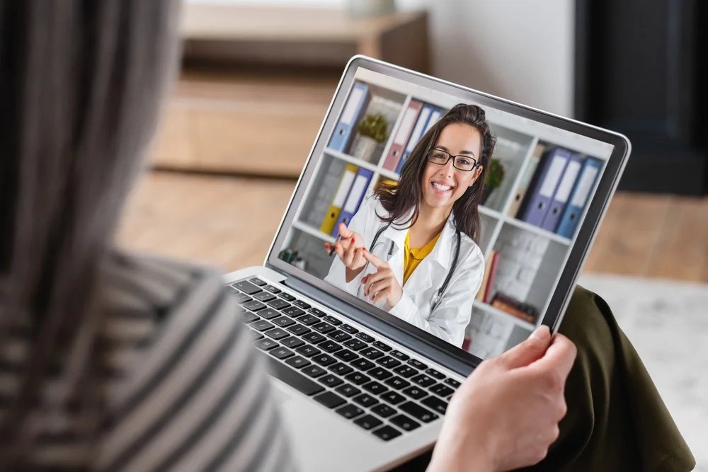 Person on video call with a female doctor with long dark hair, wearing glasses and a white coat, smiling and holding a pen, in front of bookshelf with binders.