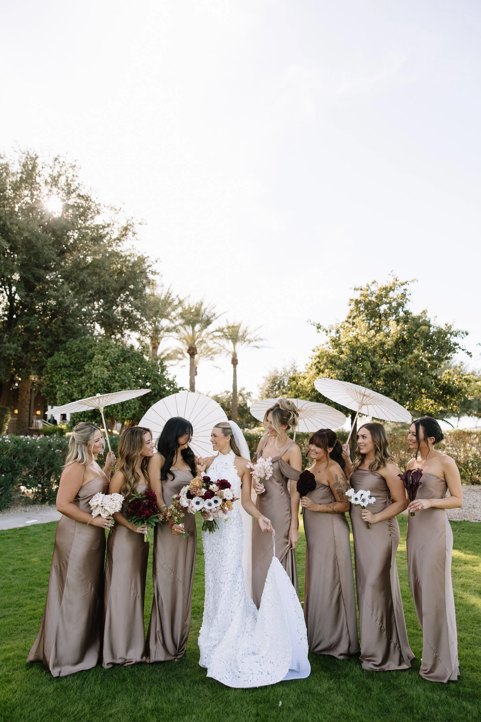 A bride in a white lace wedding gown and her bridesmaids in taupe dresses are standing on grass, holding flowers and white parasols, with trees and palm trees in the background during an outdoor wedding.