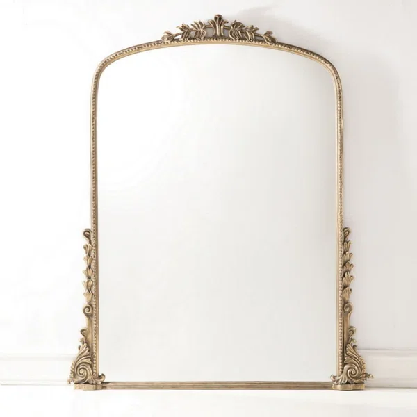 An ornate gold-framed mirror with decorative floral and scroll details at the top and bottom, mounted against a white wall.
