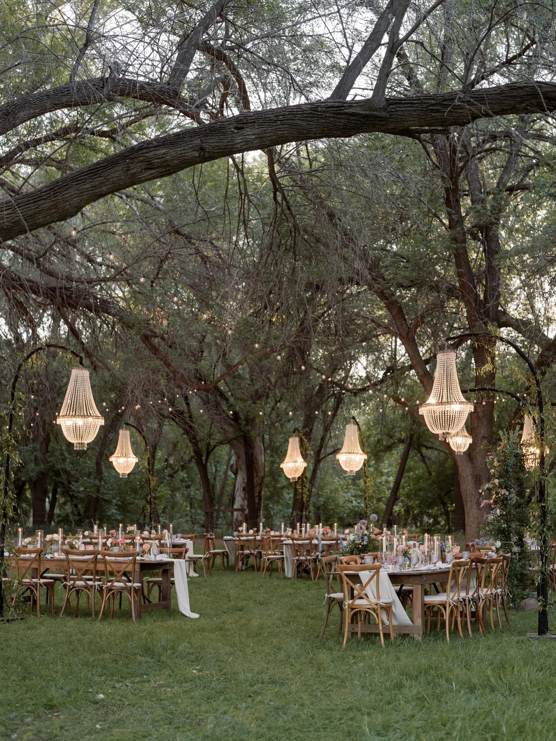 Elegant outdoor dining setup with wooden tables, chairs, floral centerpieces, candles, and hanging chandeliers in a forested area at dusk.