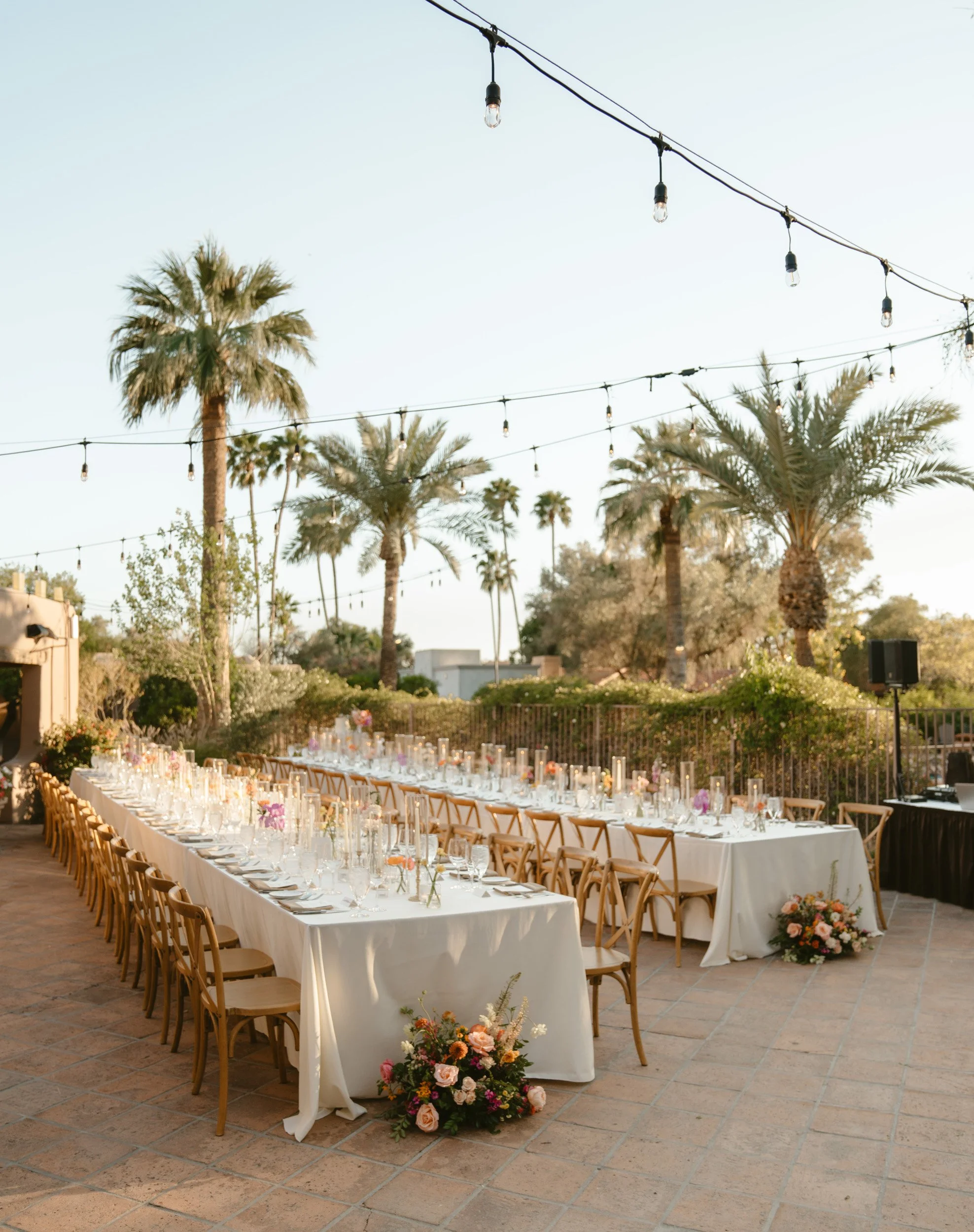 Outdoor elegant dining setup with long white tablecloth-covered tables, floral centerpieces, and wooden chairs, decorated with string lights and palm trees in the background.