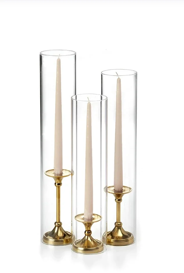 Three tall glass candle holders with gold bases, each containing a white candle, arranged in front of a white background.