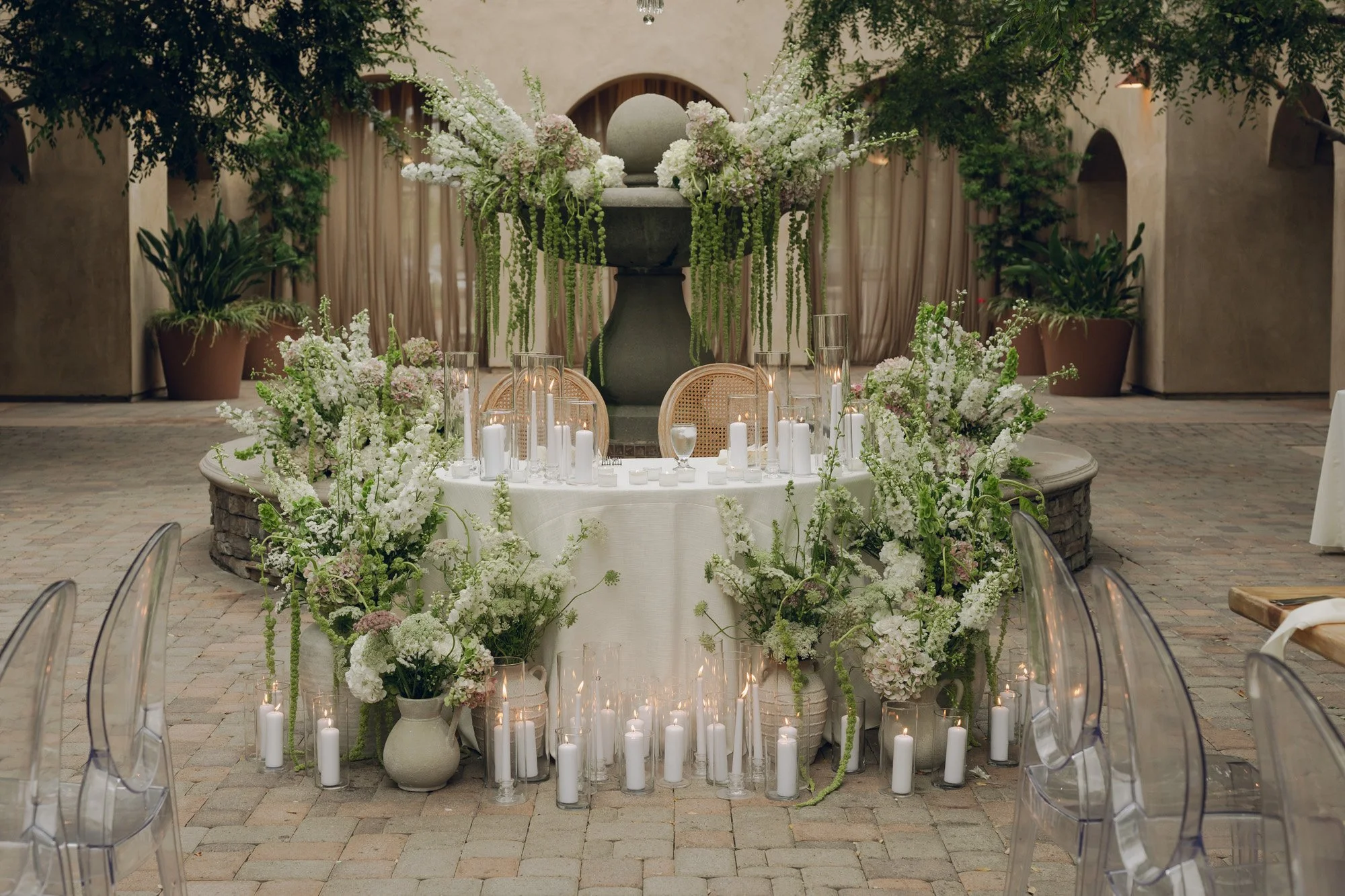 Elegant table setting with white flowers, candles, and a large floral arrangement in a courtyard with large potted plants.