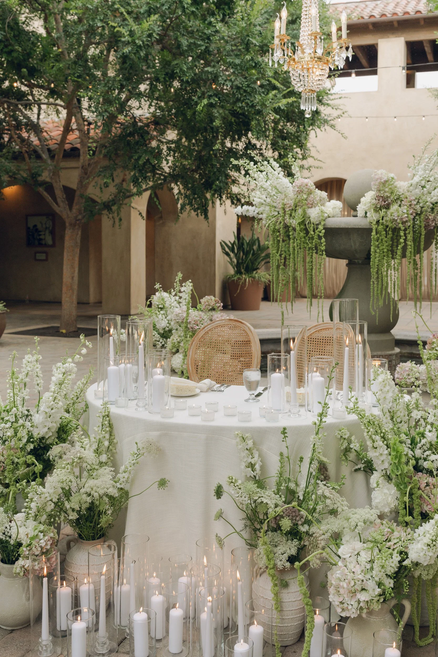 A decorated outdoor event space with a round table covered with a white tablecloth, surrounded by flowers and candles. The table features tall glass candle holders with white candles, and the surrounding area has white flowers in pots and vases, creating an elegant and lush setting. There is a large floral arrangement on a stone pedestal and a chandelier hanging above.