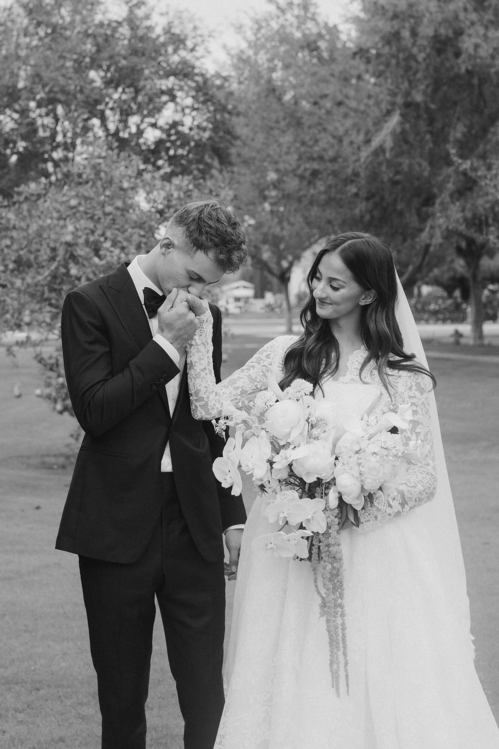 A groom kisses a bride's hand while holding her hand, with the bride smiling and holding a large bouquet of flowers, outdoors in a park with trees in the background.