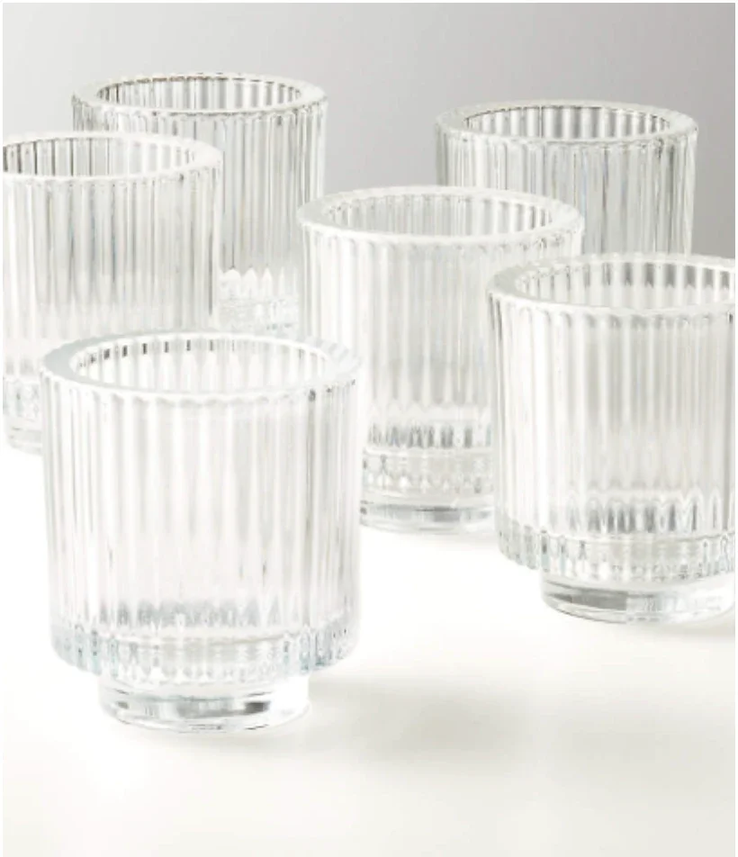 Six clear glass drinking cups with vertical ridges, arranged on a white surface against a light background.