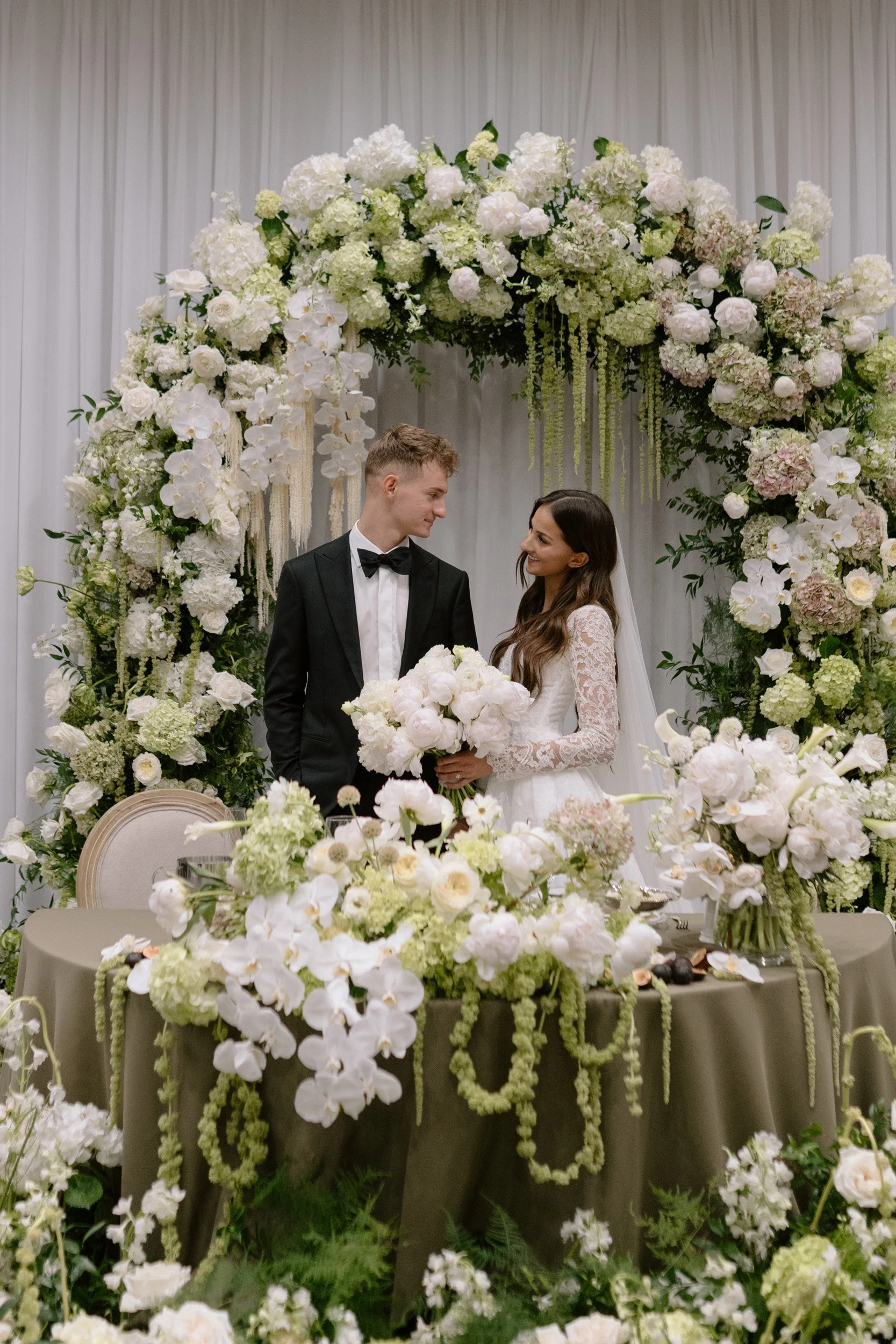A bride and groom standing face to face at their wedding, surrounded by a white floral arch with white flowers and greenery, with a table decorated with matching flowers in front of them.