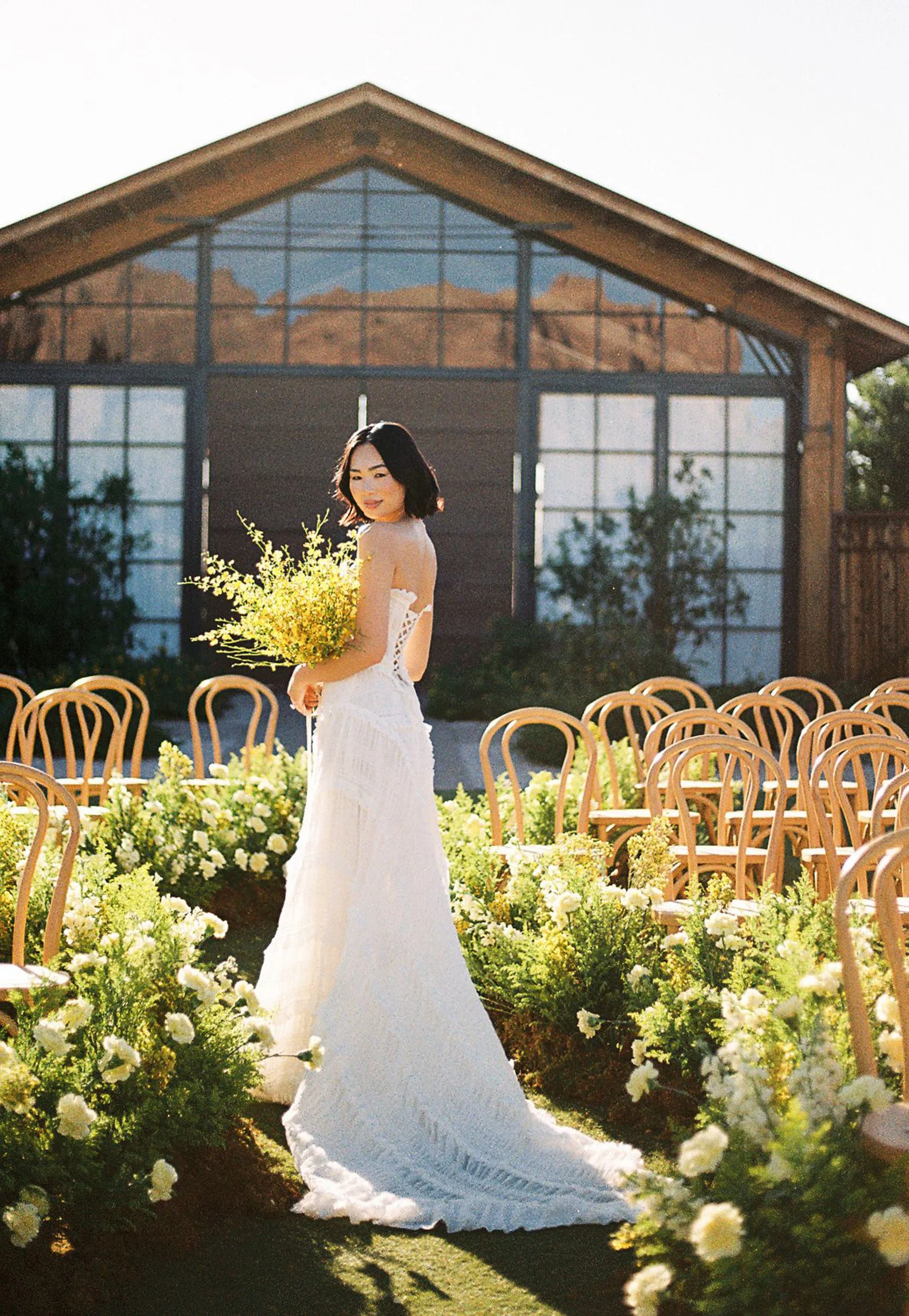 A woman in a white wedding dress holding a bouquet of yellow flowers, standing in an outdoor wedding ceremony setting with chairs and floral arrangements, in front of a modern wooden and glass building.