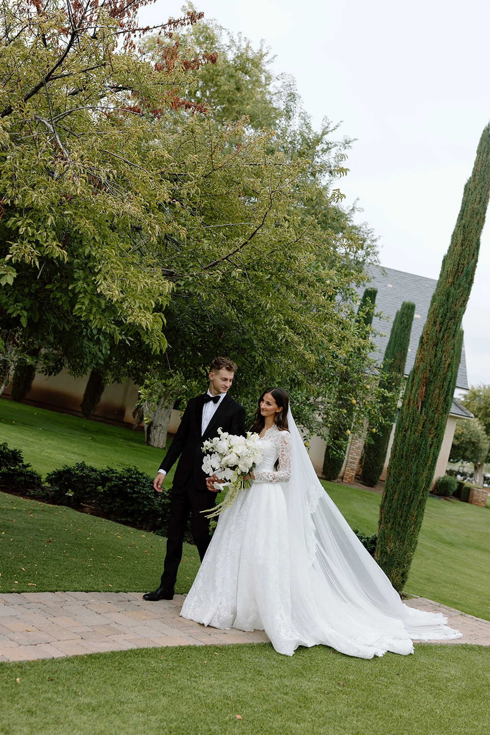 A bride and groom walking in a garden on their wedding day, with the bride holding a bouquet of white flowers and wearing a long white lace wedding dress with a veil, and the groom in a black tuxedo with a bow tie, surrounded by green trees and neatly trimmed grass.