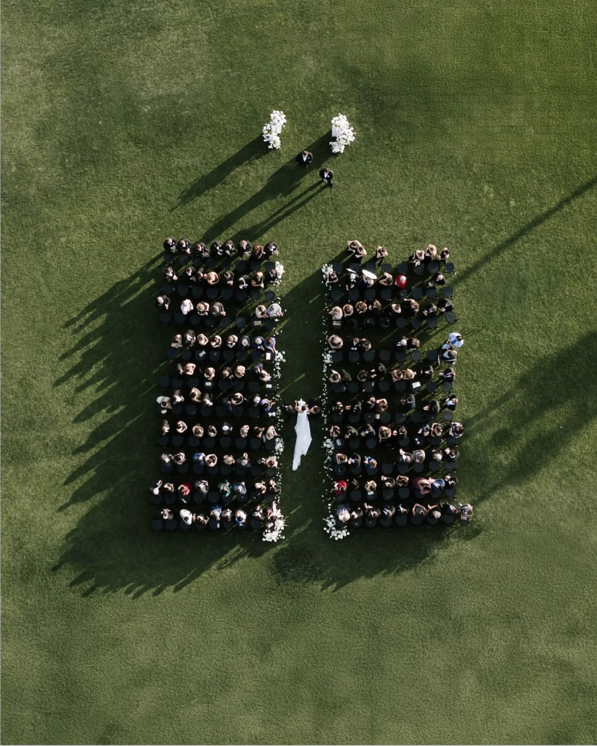 Aerial view of a wedding ceremony with guests seating in rows on green grass, and a bride and groom standing at the front near flower arrangements.