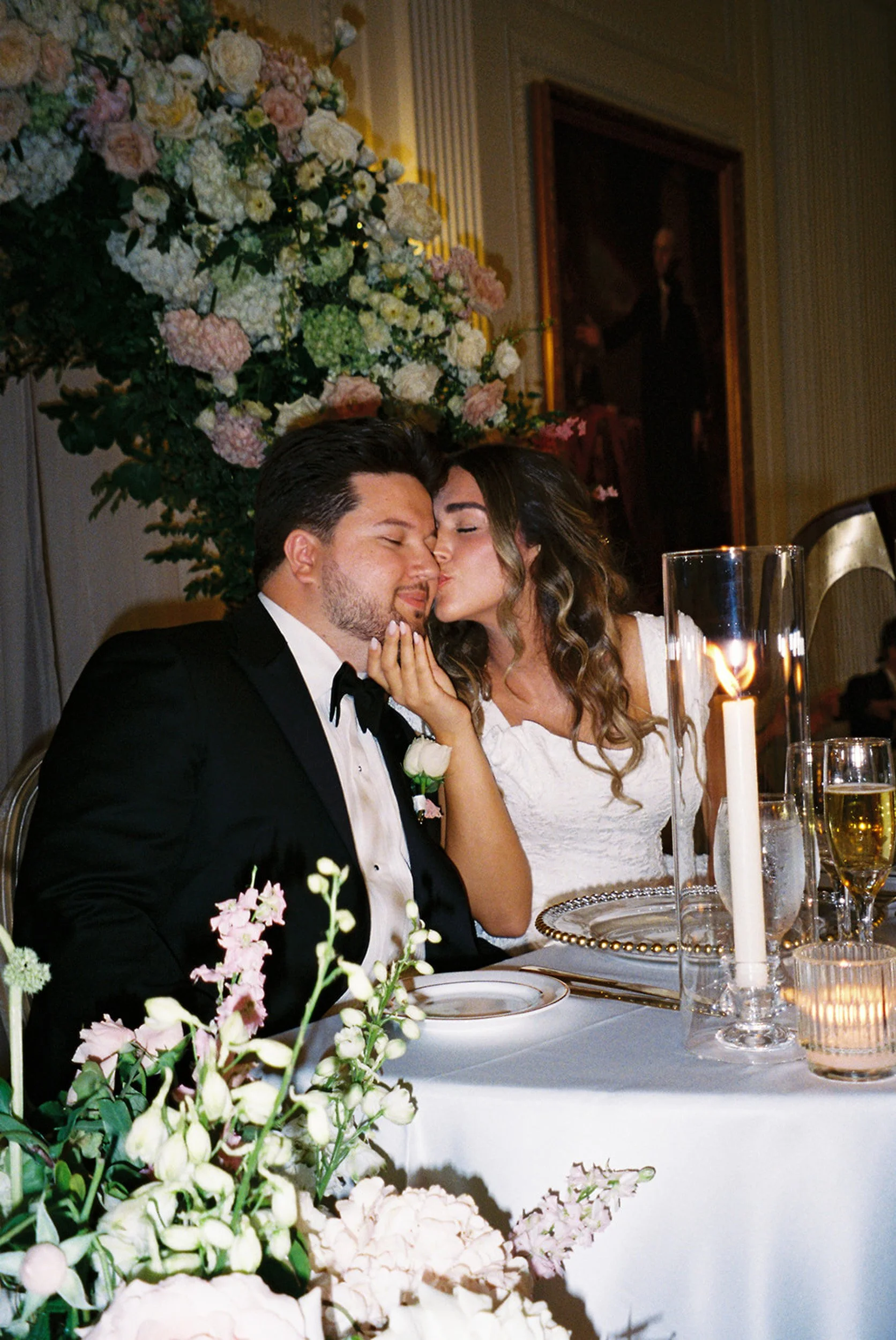 A bride and groom share a kiss at their wedding reception, sitting at a decorated table with flowers, candles, and champagne.