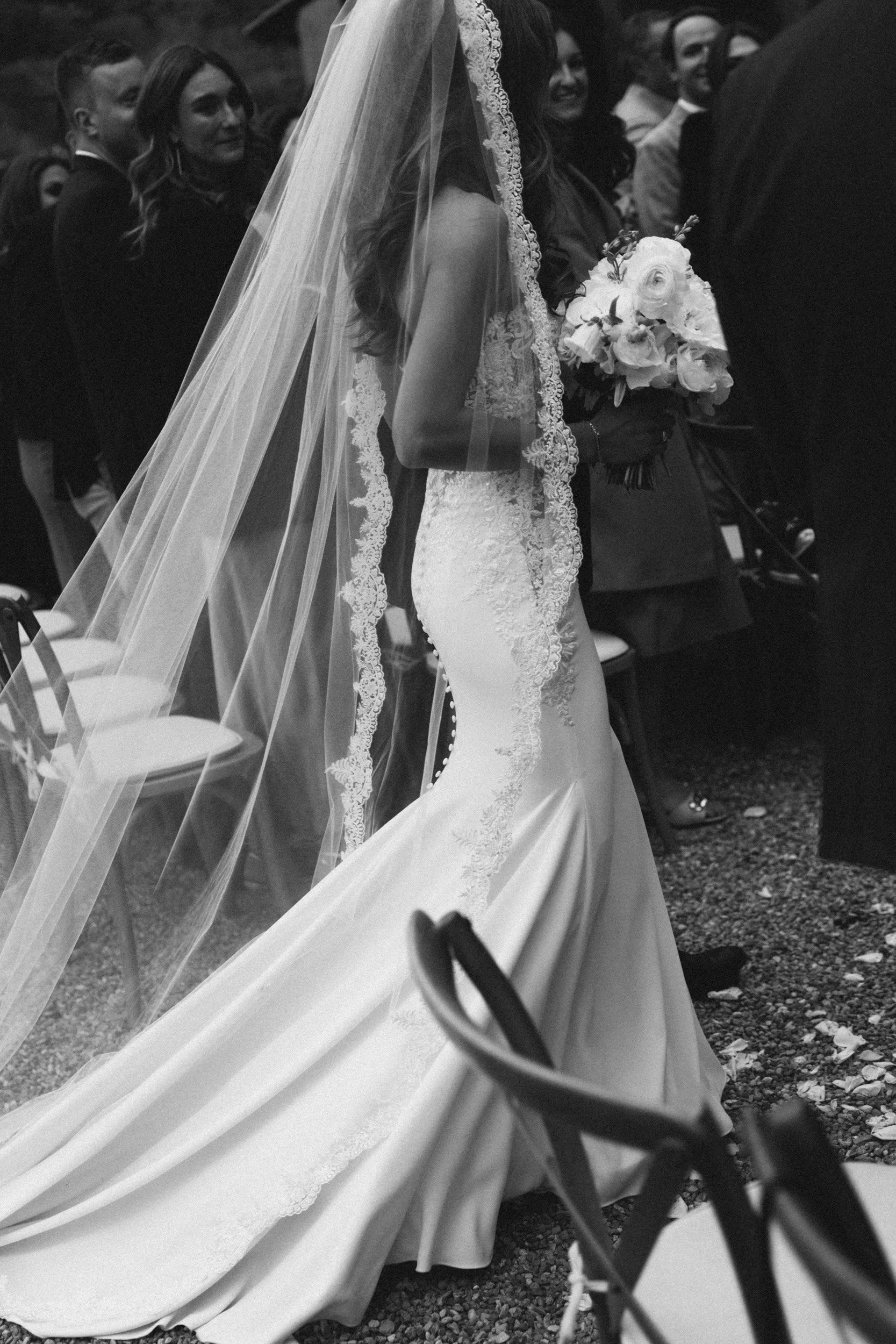 A bride in a wedding dress and veil holding a bouquet, surrounded by seated guests at an outdoor wedding ceremony.
