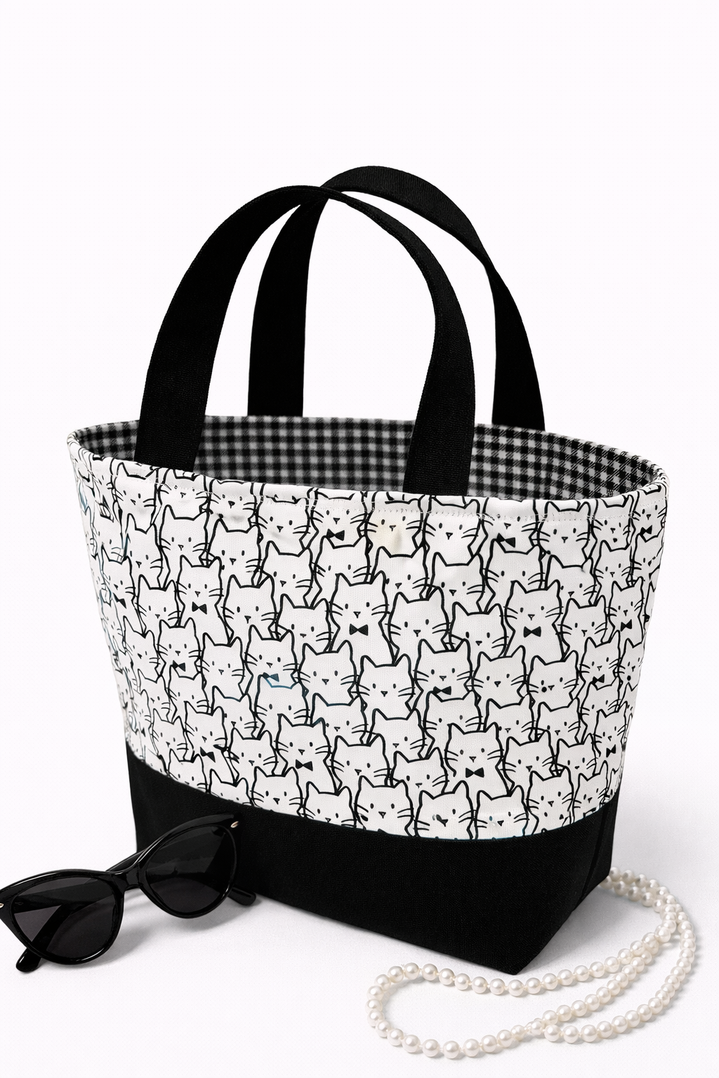 A tote bag with a cat pattern, a string of pearls, and a pair of black sunglasses placed on a white surface.