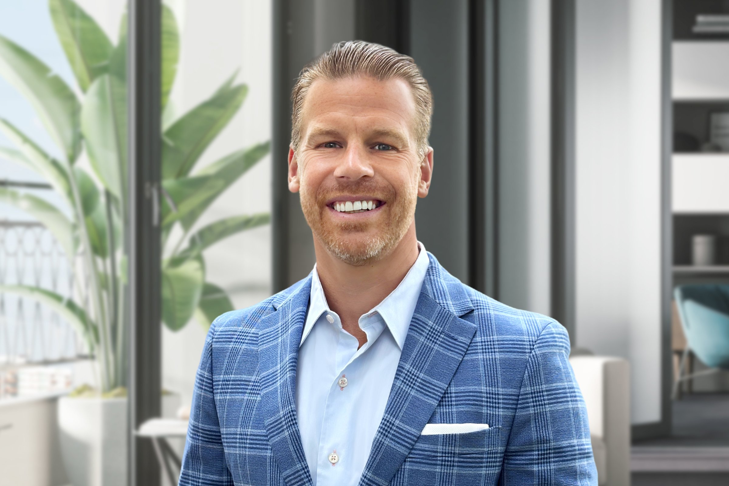A smiling man in a blue plaid suit jacket and light blue shirt standing in a modern office with large windows and green plants.