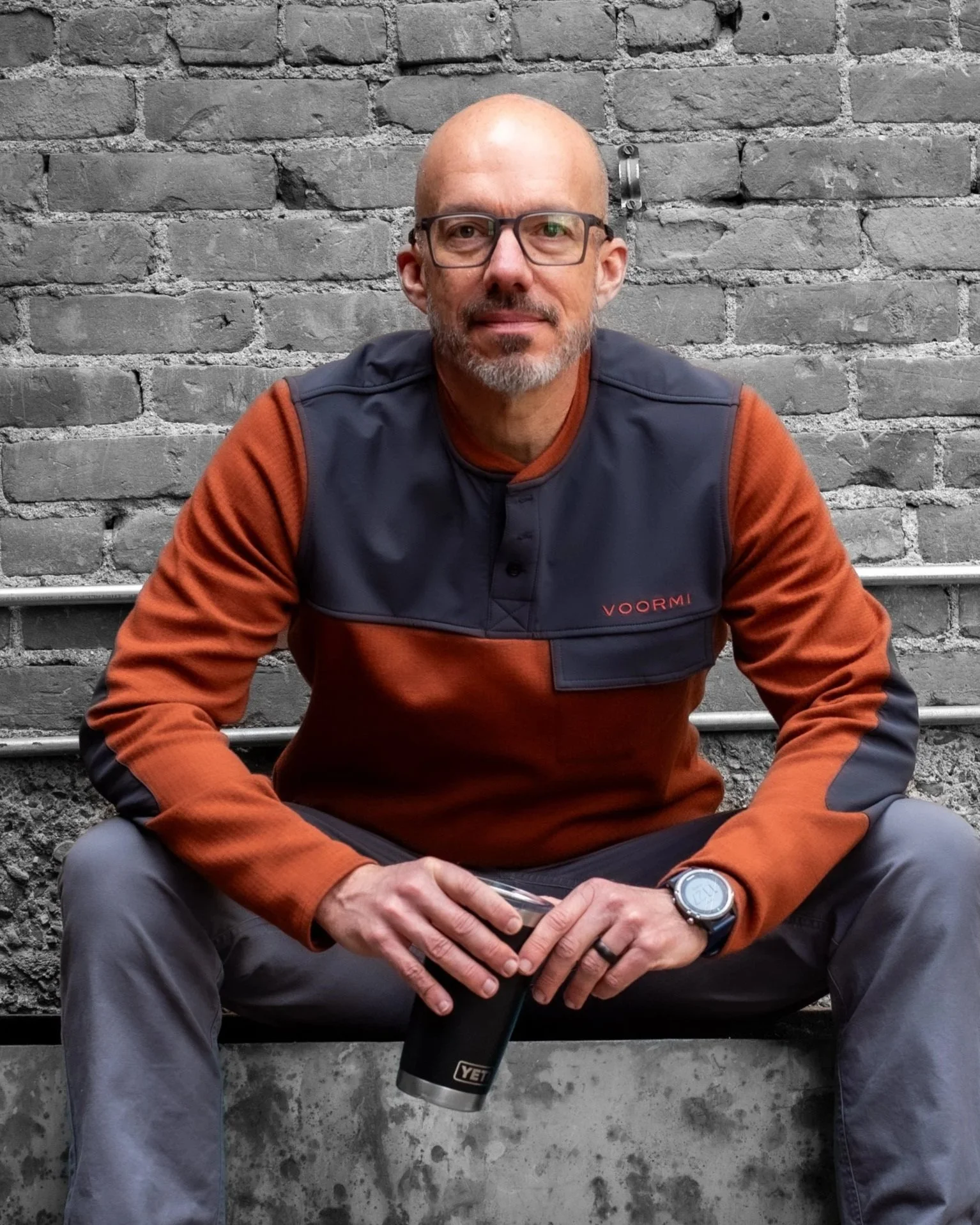 A bald man with glasses and a beard sitting on concrete stairs against a gray brick wall, holding a black YETI travel mug, wearing a brown and black performance jacket with the word "VOORMI" on it, and a watch on his left wrist.