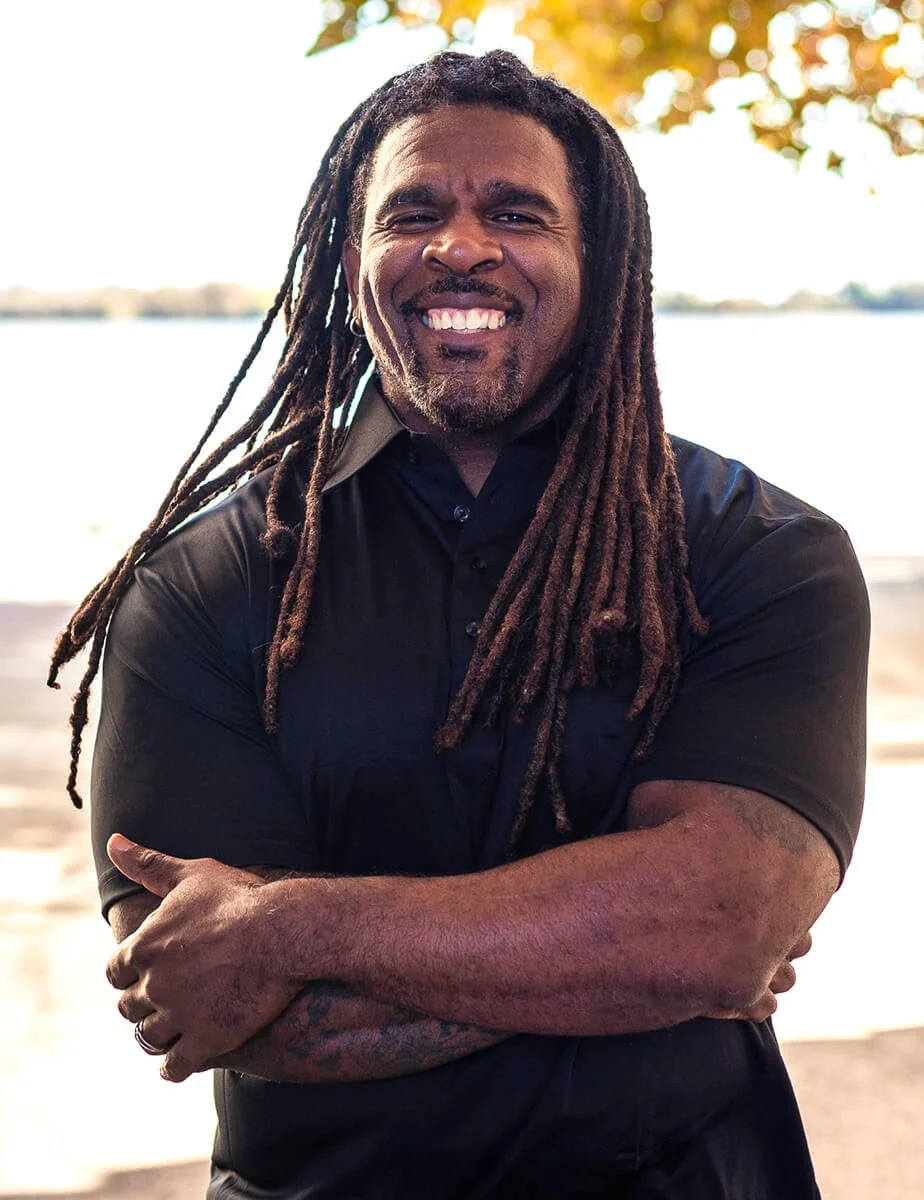 A man with long dreadlocks smiling outdoors near a body of water, with trees and sunlight in the background.