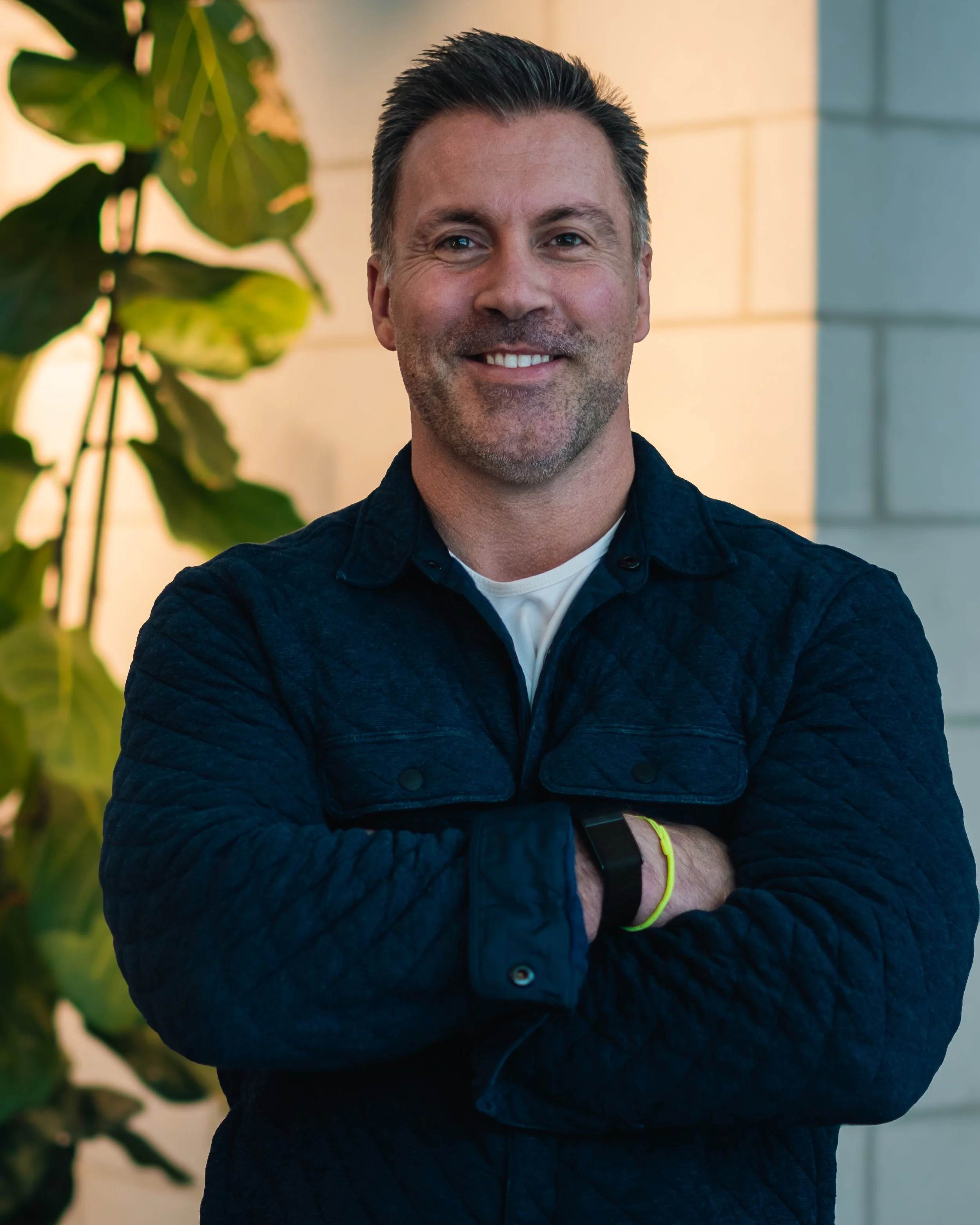 A man with short dark hair and a light beard, smiling, standing with arms crossed in front of a light-colored brick wall and green leafy plant.
