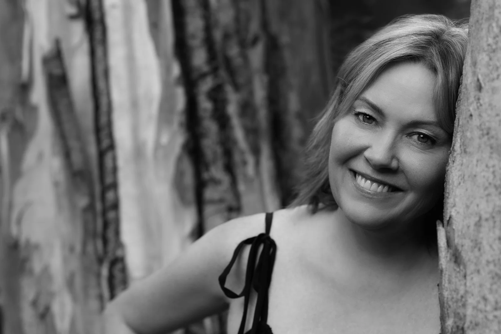 A black and white portrait of Colleen Dolan, leaning against a textured tree trunk outdoors.