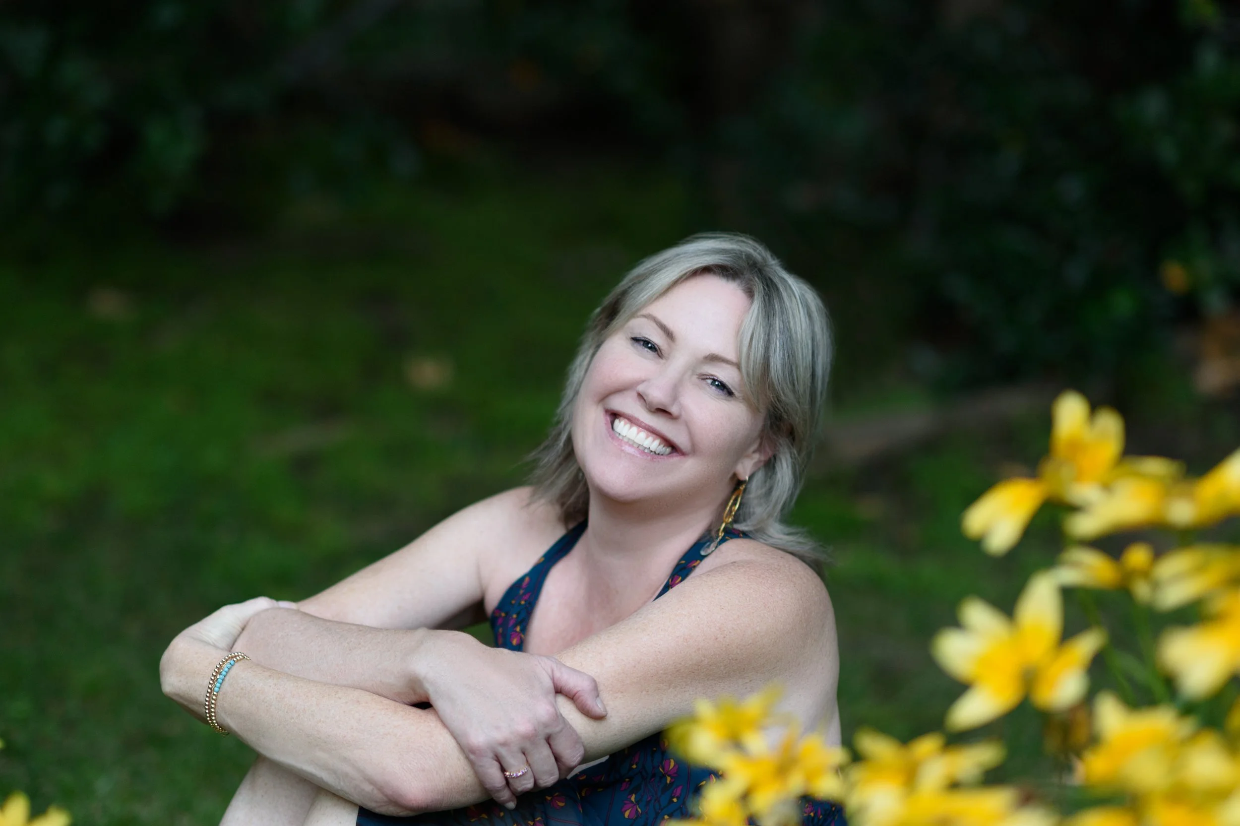 Colleen Dolan smiling outdoors with green foliage and yellow flowers in the background.