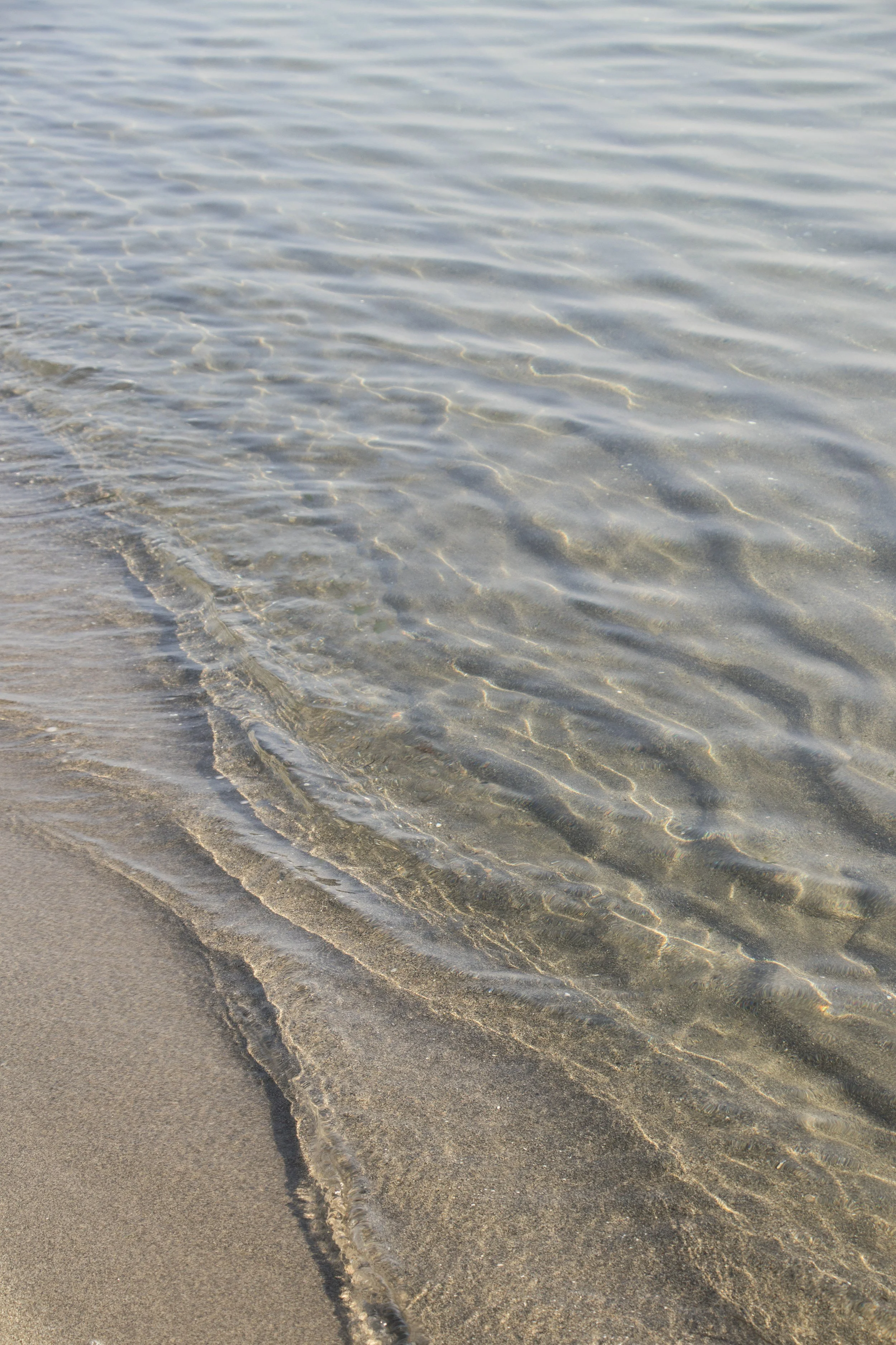 Close-up of a sandy beach with gentle waves of water reaching the shore, creating ripples in the shallow, clear water.