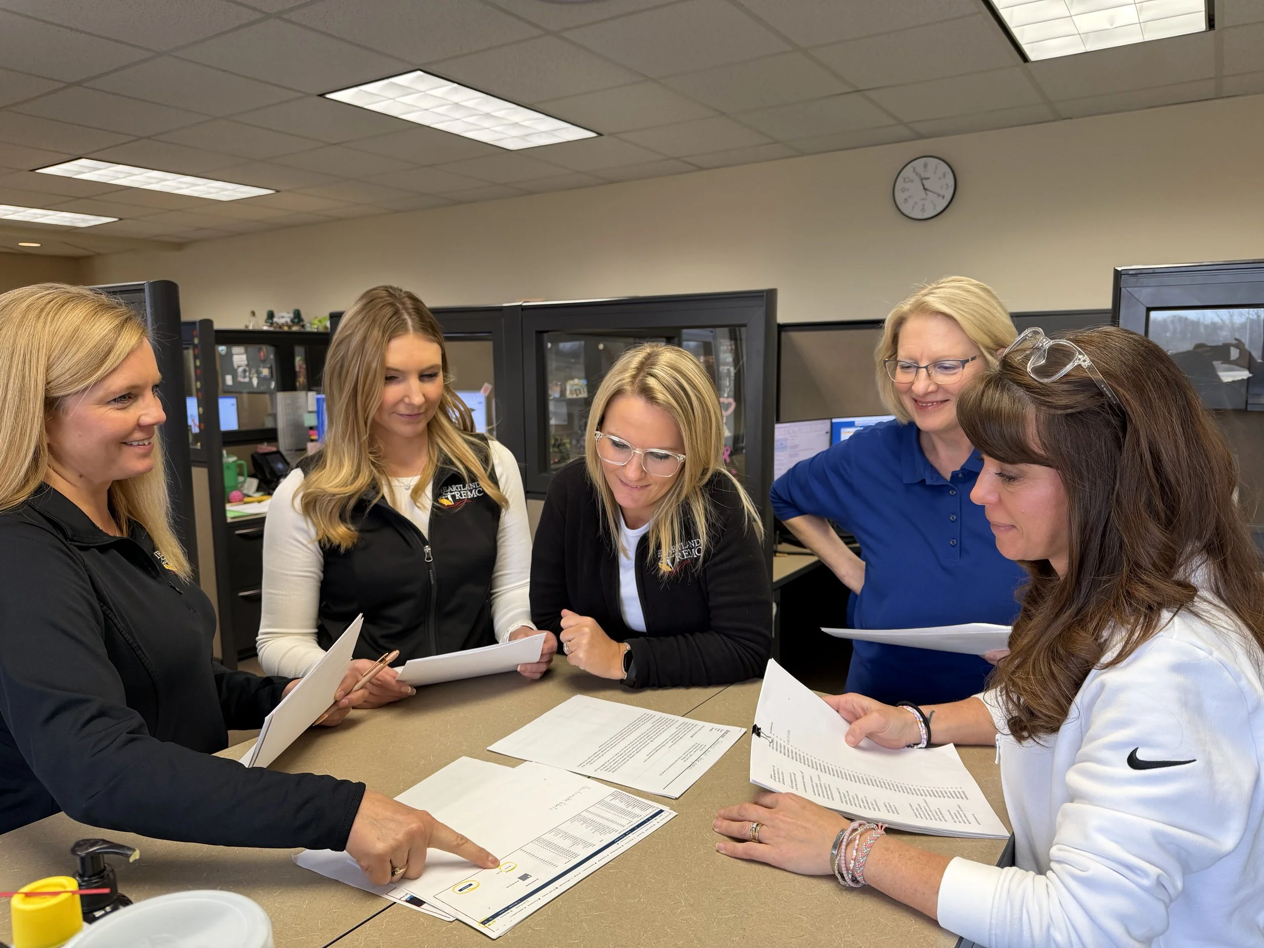 A group of women gathered around a table in an office, reviewing documents together. They are smiling and engaged in discussion, with a clock on the wall showing 1:11.