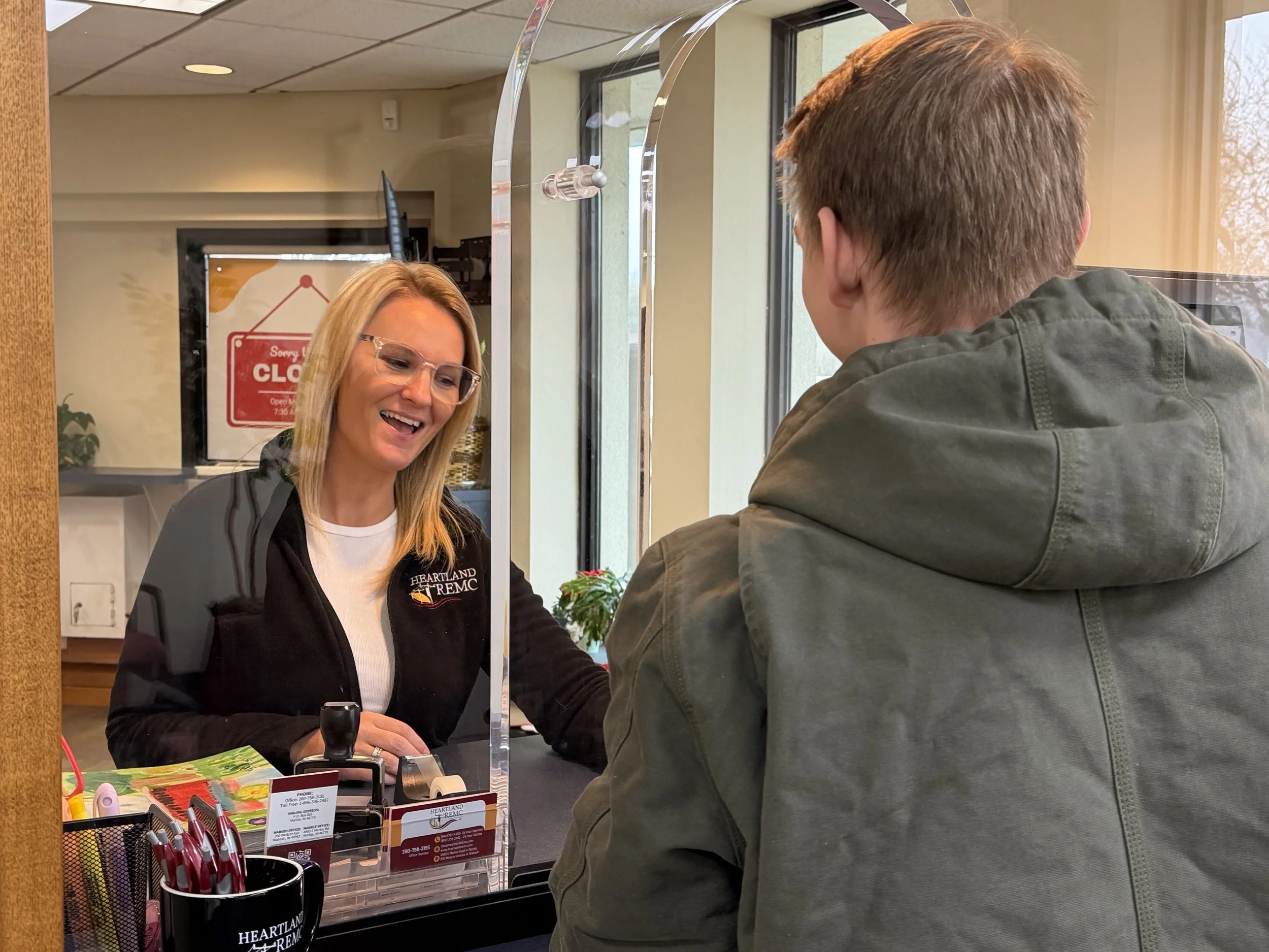 A woman with blonde hair and glasses smiling at a man across a glass partition in a reception area, as he pays his bill.