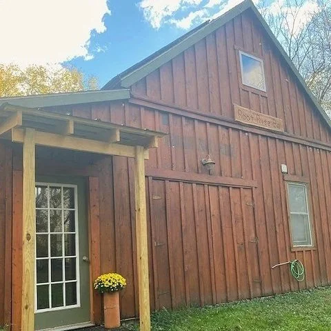 A wooden barn-style building with a sign that reads 'Root Rise Farm,' a potted yellow flower near a glass-paneled door, and a garden hose on the wall.