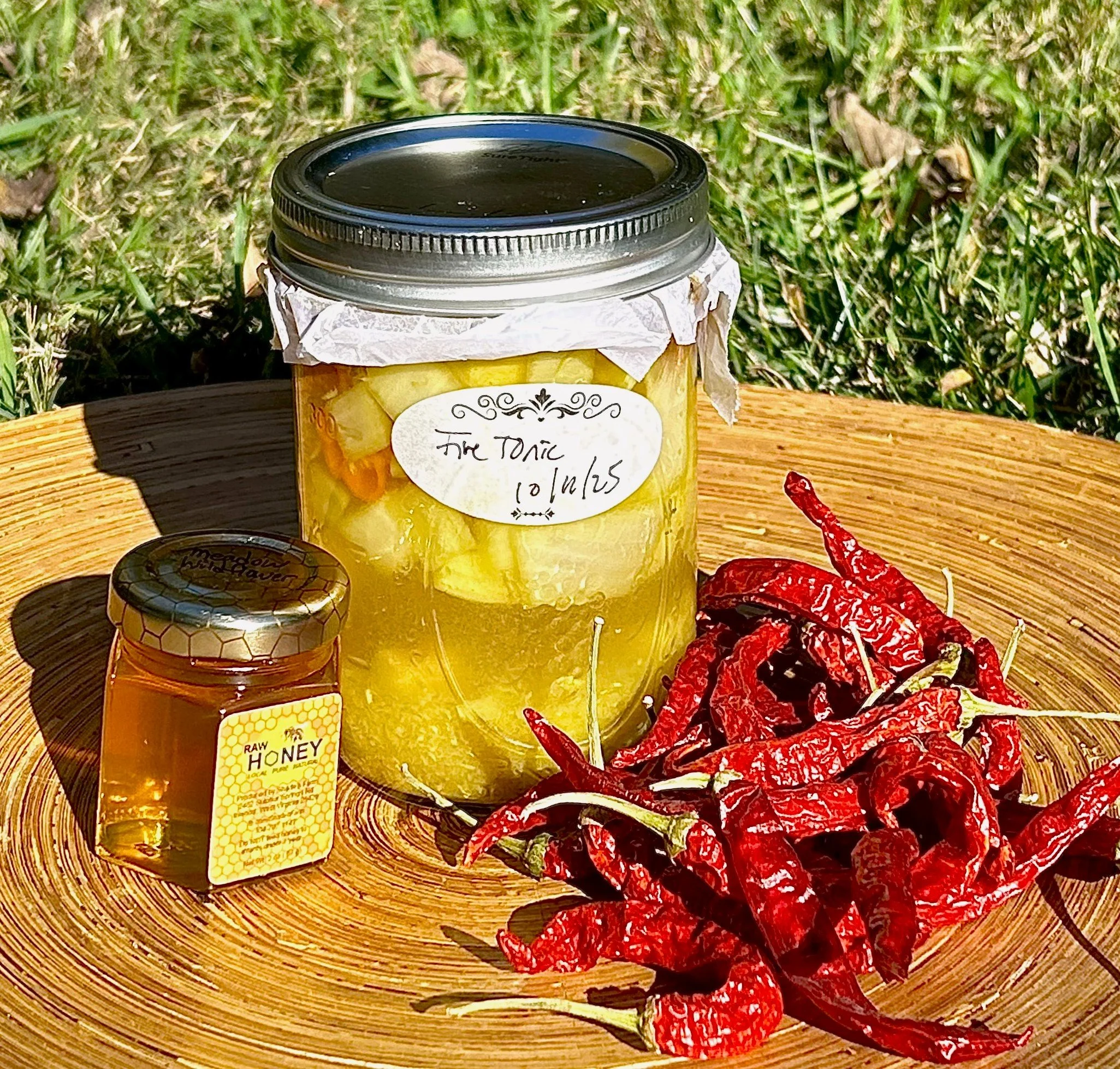 A jar of sliced pineapple labeled 'The Tonic,' a small jar of honey labeled 'Raw Honey,' and a bunch of dried red chili peppers on a round wooden plate outdoors with grass in the background.