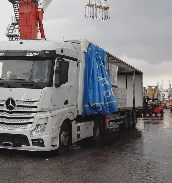 Camion Mercedes con coperchio blu parzialmente chiuso, in un'area industriale con gru e carrello elevatore, sotto un cielo nuvoloso.