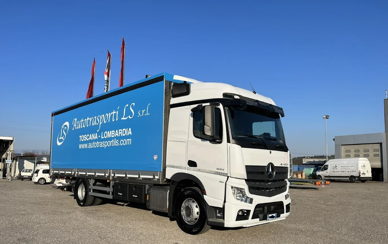 Camion Mercedes con scatola blu con scritte 'Autotrasporti LS s.r.l. Toscana - Lombardia', in un'area industriale con altri veicoli e un edificio.
