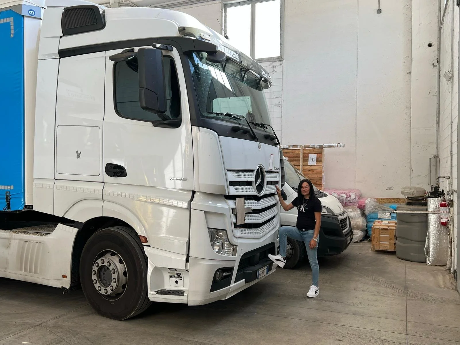 Una donna in piedi accanto a un camion Mercedes-Benz bianco all'interno di un magazzino, con vari oggetti e pallet sullo sfondo.