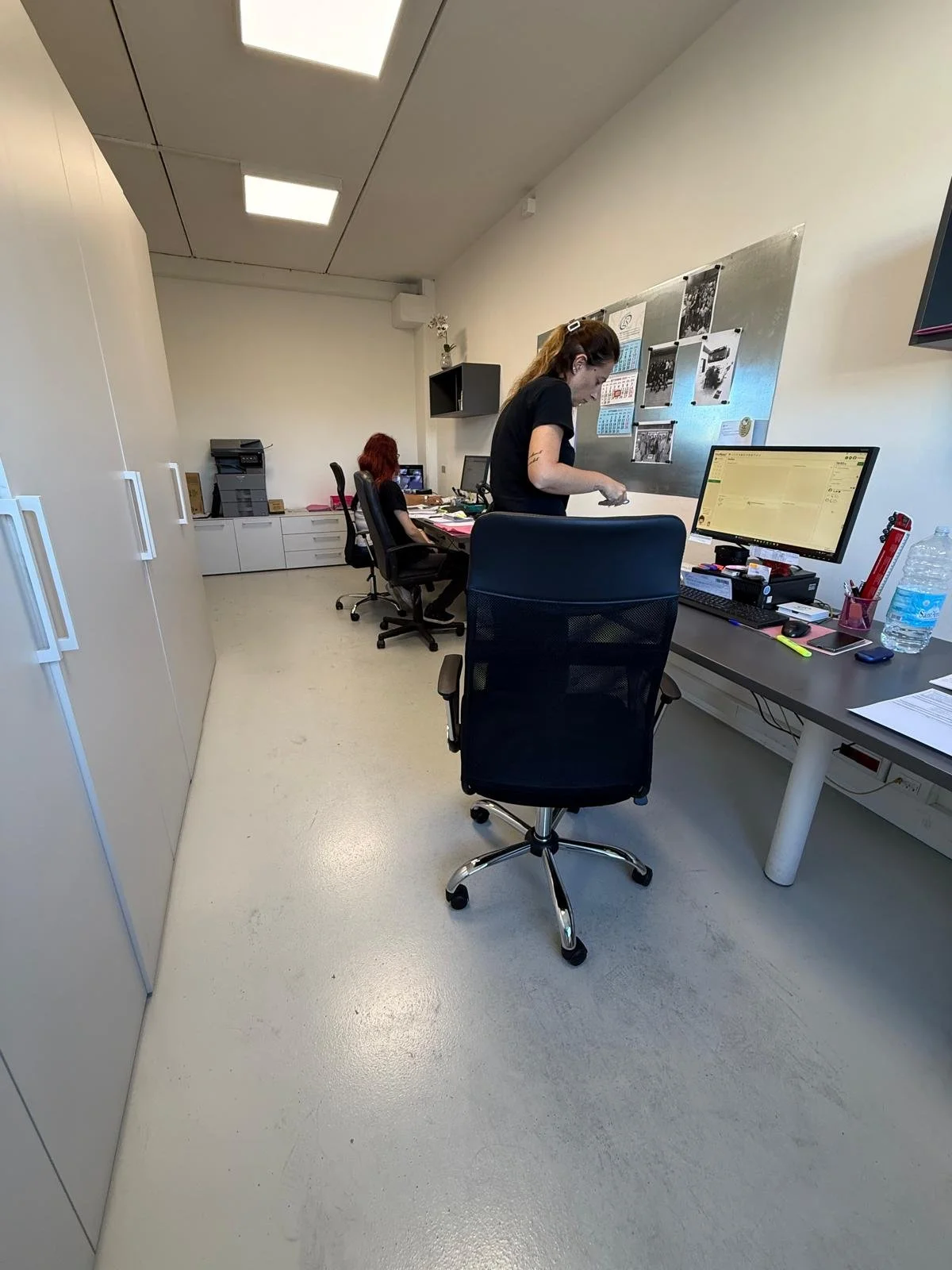 Ufficio con tre donne che lavorano ai computer. Una donna è in piedi mentre le altre sono sedute. Presente una lavagna con foto e documenti, e una grande bottiglia d'acqua sul tavolo.