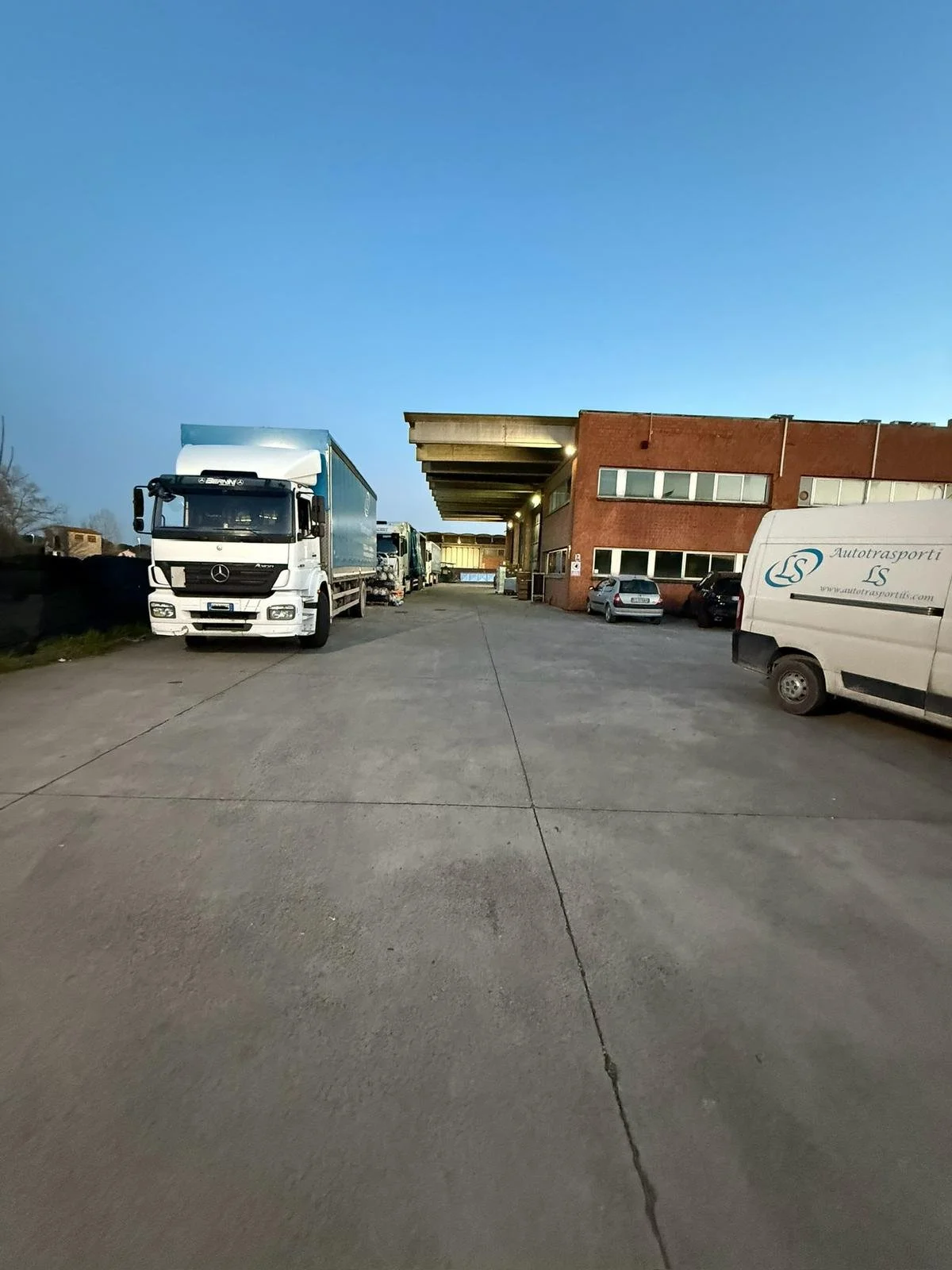 Area di carico con diversi camion e veicoli parcheggiati di fronte a un edificio industriale vecchio, durante il crepuscolo o la sera.